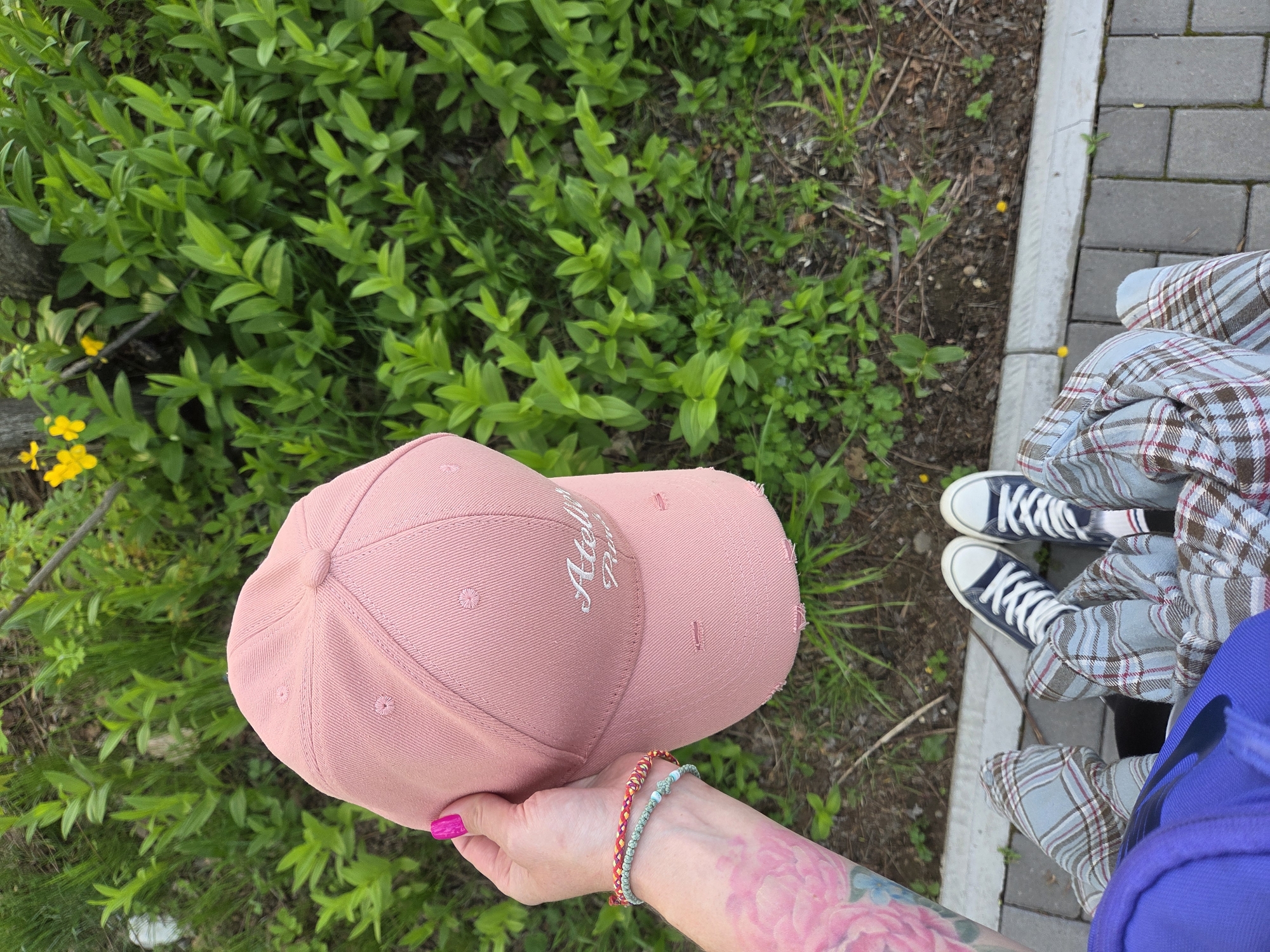 A person is holding a pink baseball cap with white writing in their left hand. The person has pink nail polish and two bracelets on their left wrist. They also have a tattoo of a pink flower on their left arm. The background includes green bushes, yellow flowers, pavement bricks, and a person wearing blue sneakers with a plaid pattern on their pants. The location is outdoors and likely near a city street. Birobidzhan, Russia, is not discernible from the image.