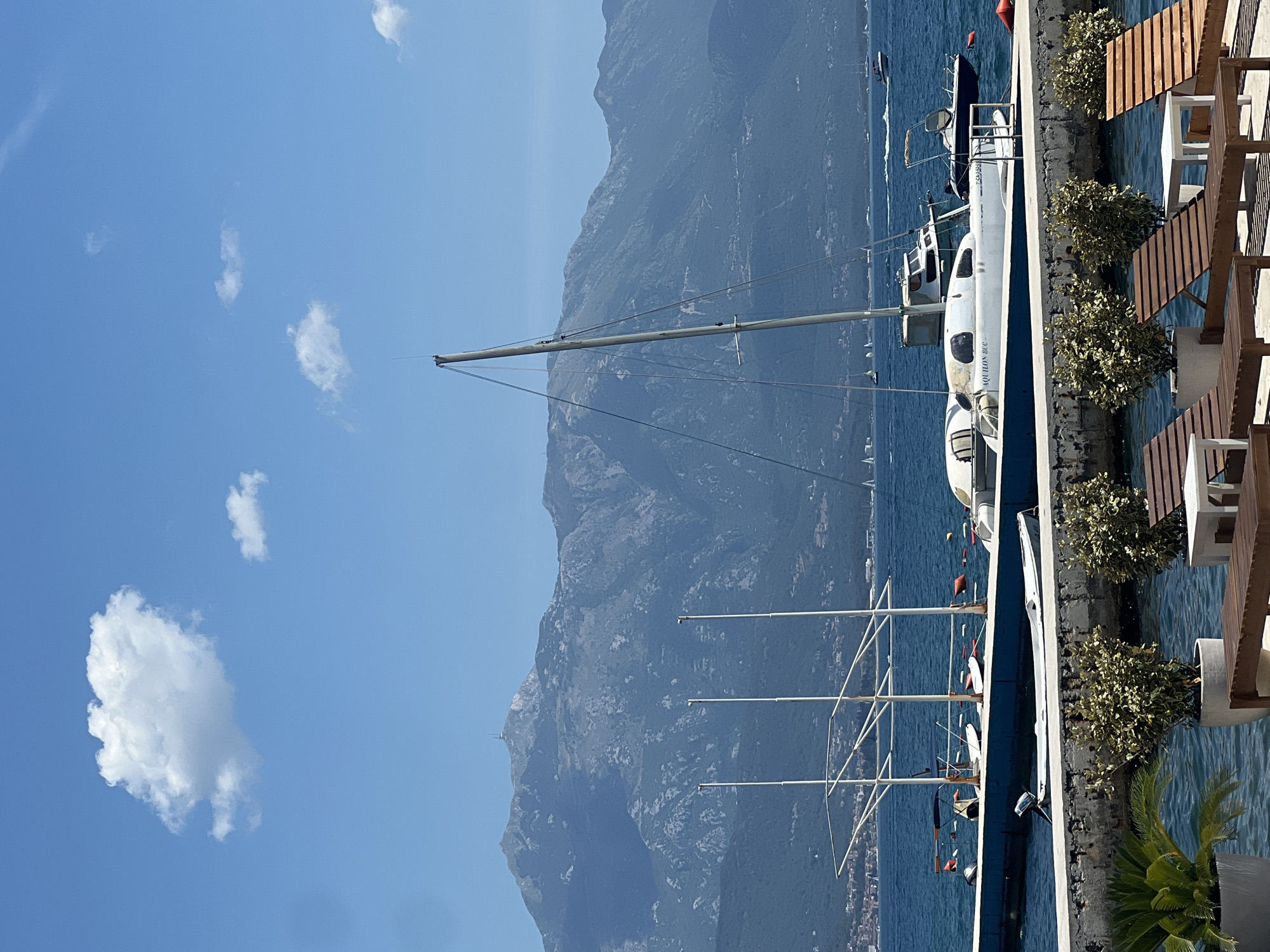 A bright, sunny day unfolds over the Bay of Kotor in Baošići, Montenegro. The foreground features a paved waterfront promenade lined with potted plants and wooden lounge chairs, indicating a relaxed seaside setting. Beyond the promenade, the deep blue waters of the bay are dotted with various boats, including a prominent white catamaran with the name "AQUILON 8CC" visible on its hull. Several other vessels, ranging from small motorboats to sailboats, are scattered across the water.
In the mid-ground, a concrete pier juts out into the bay, supporting several white poles, possibly remnants of a former structure, and a series of buoys. Moored to this pier are more boats, including a small white tender and the aforementioned catamaran. Further out on the water, a white motorboat speeds across the bay, leaving a wake.
The background is dominated by the majestic, rugged mountains that frame the Bay of Kotor. Their slopes are covered in a mixture of dark green vegetation and lighter rocky outcrops. A prominent peak in the center of the mountain range features a small structure near its summit, likely a communication tower. The sky is a clear, vibrant blue, punctuated by a few fluffy white clouds. The overall impression is one of a peaceful, scenic coastal location.