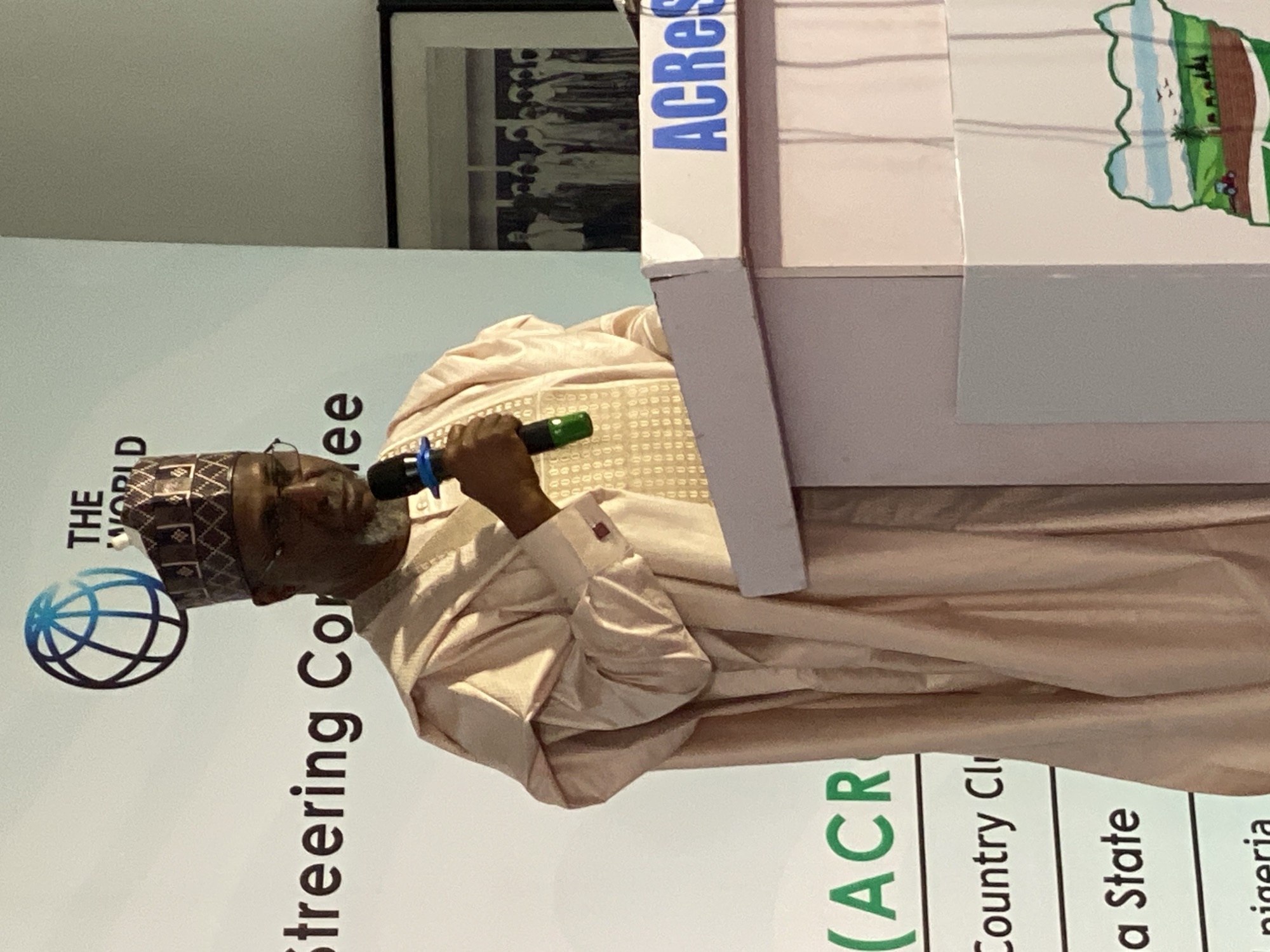 A man is speaking at a podium. He is wearing a patterned cap and light pink robes. He is holding a microphone with a blue and green handle. The podium has the word "ACRes" written on it vertically in blue letters. The background behind the man contains the text "THE WORLD Steering Committee" and a globe icon. The text "(ACR Country Cli a State nigeria" is also visible near the bottom of the image. A framed picture hangs on the wall behind the podium. The location is identified as Kwarau, Nigeria.