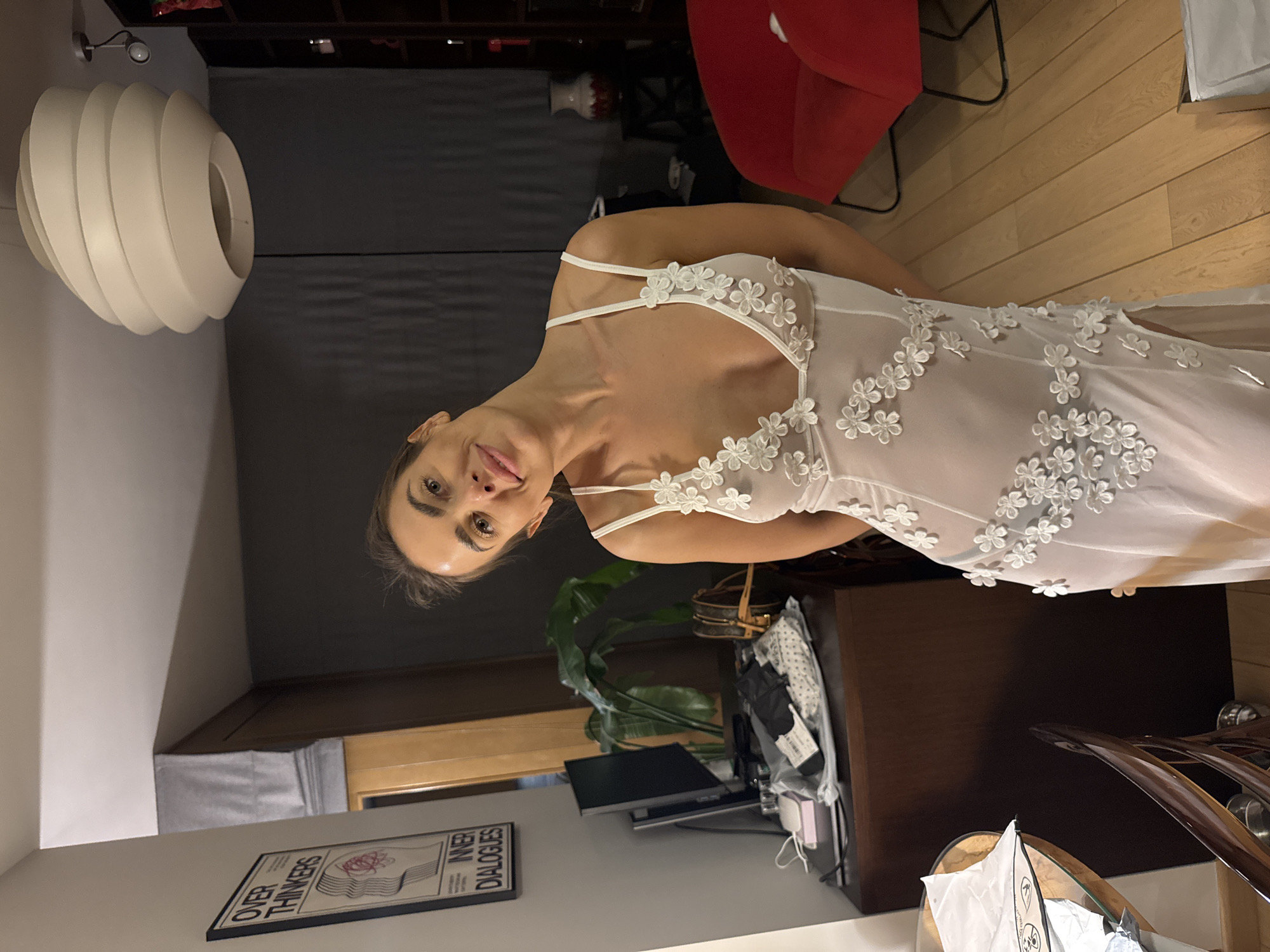 A woman in a white floral dress stands indoors, leaning into the frame. Behind her, a room includes a red chair, a cabinet, a lamp hanging from the ceiling, and a poster labeled "OVER THINKERS INNER DIALOGUES" on the wall. A houseplant sits near a desk with a computer monitor. The location may be in Warsaw, Poland.