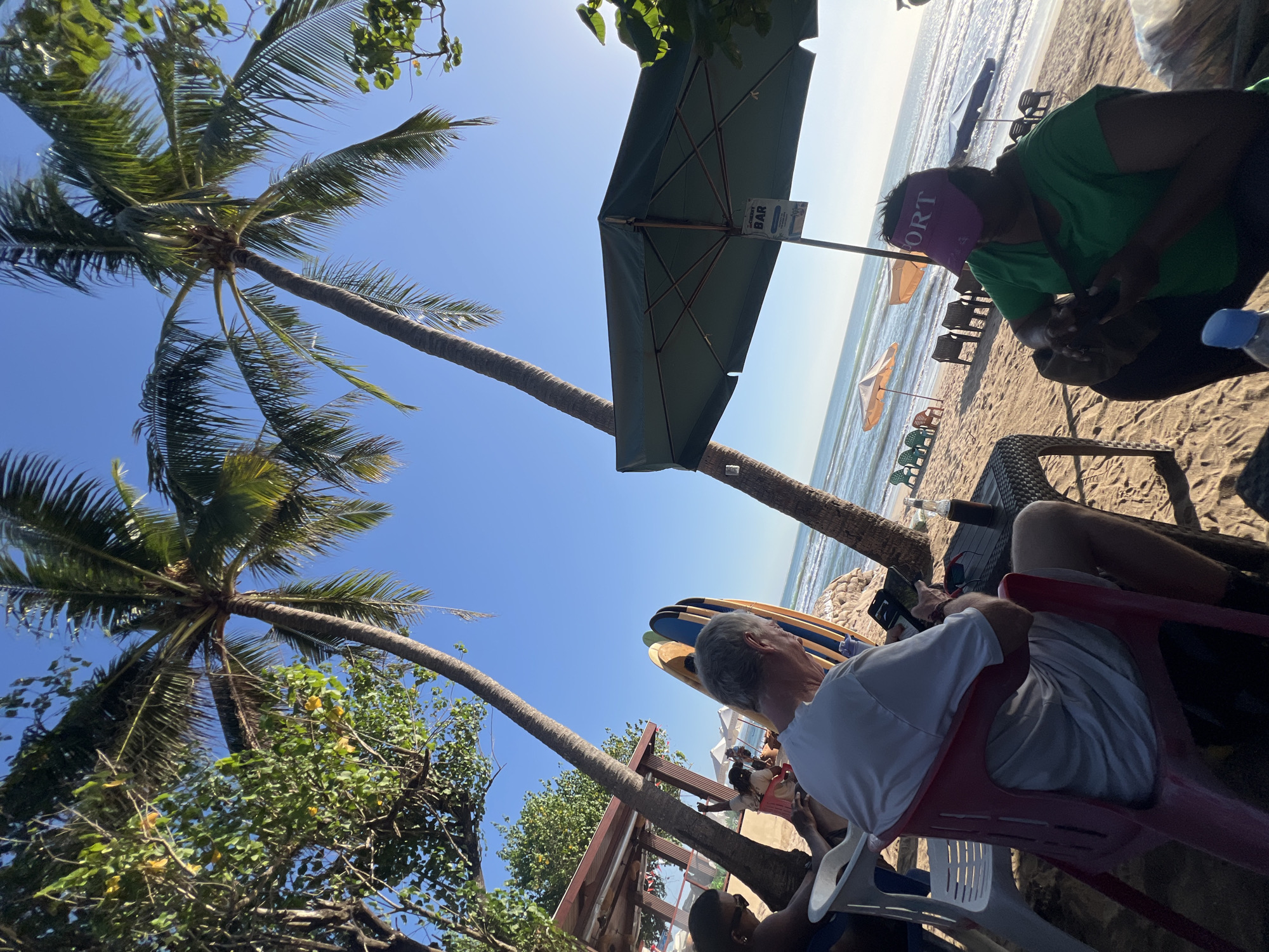 The image depicts a beach scene in Kuta, Indonesia, likely a resort area. In the foreground, a man in a white t-shirt and shorts sits in a red plastic chair at a woven table, looking at his phone. Beside him is a bottle of beer. To his right, a woman in a green t-shirt and a pink visor that reads "SPORT" is looking down at something in her hands. She has a bag slung across her chest. Behind them, surfboards are propped up against a rocky embankment. Palm trees are prominent in the scene, with their fronds extending across the bright blue sky. In the background, beach chairs and umbrellas are scattered on the sandy shore, and the ocean with gentle waves is visible. A structure with a red roof, possibly a bar or restaurant, is partially visible on the left side of the image. The sun appears to be low in the sky, casting long shadows.