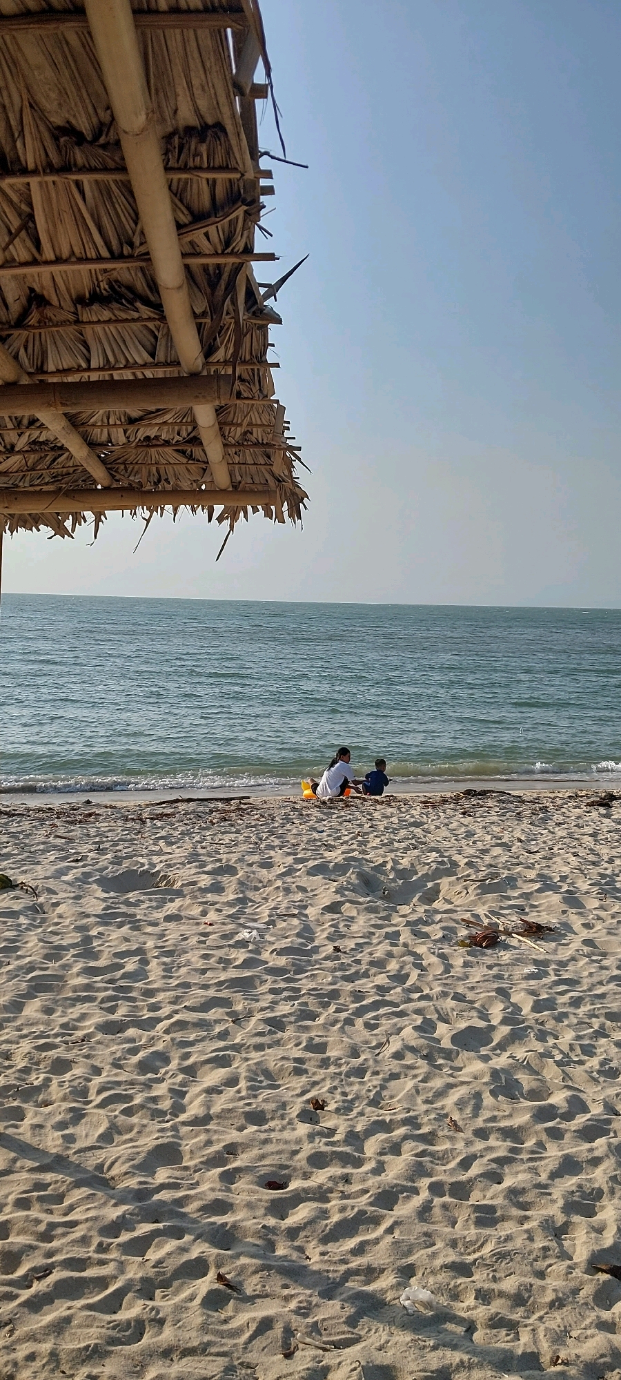 Two people, one adult and one child, are sitting on the sandy shore of a beach in Karanggondang, Indonesia. The adult is wearing a white top and the child is wearing a blue top. They are facing the ocean and appear to be playing. There is a yellow toy between them. The beach is made of light-colored sand with ripples from the tide. The ocean is a calm blue-green color with gentle waves lapping at the shore. In the foreground, part of a thatched roof structure, possibly a beach umbrella or shelter, is visible. The sky is clear and a light blue. The overall scene depicts a peaceful day at the beach.