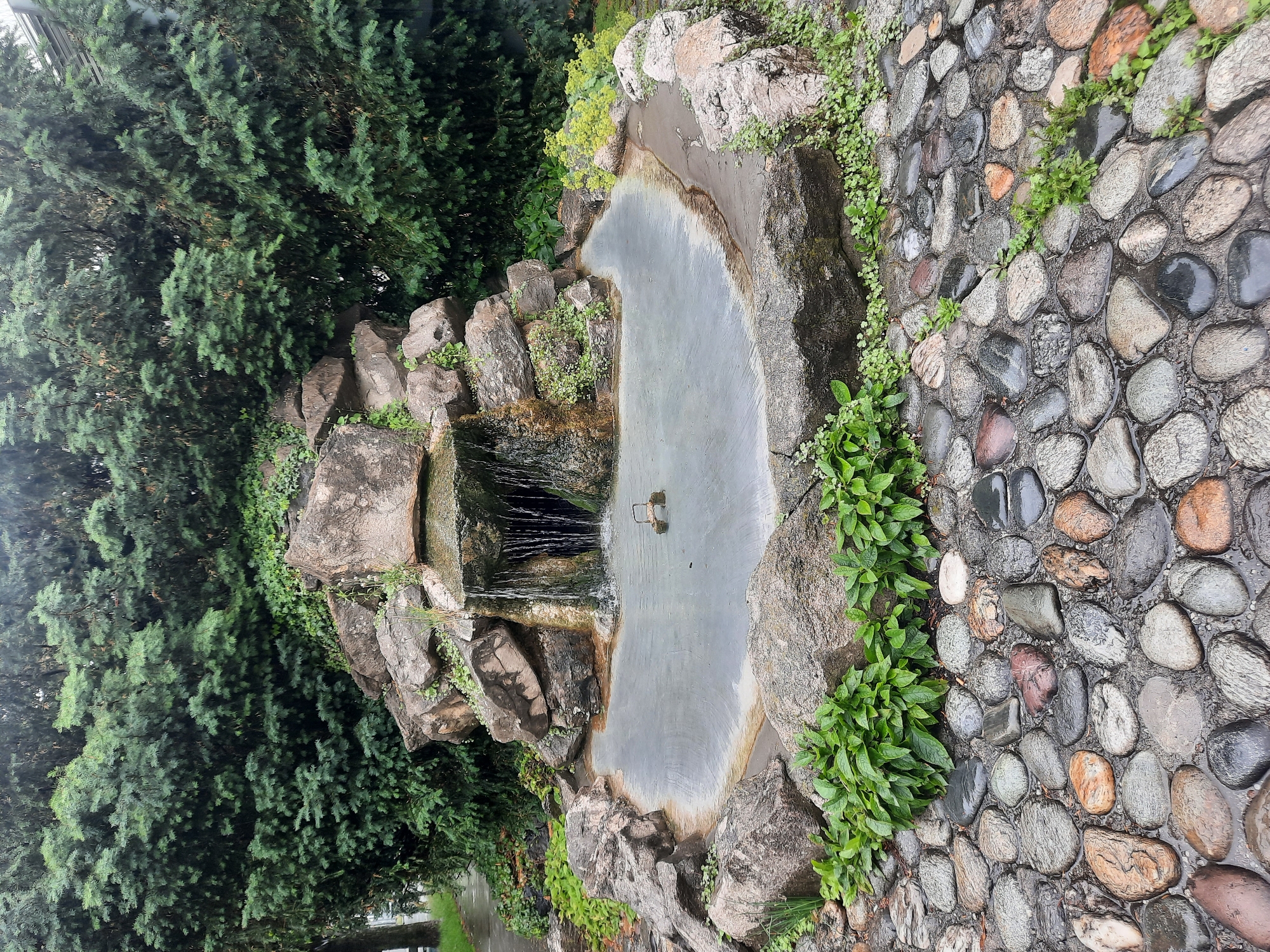 A decorative rock garden with a small waterfall and pond is shown. The garden is surrounded by lush green trees and ivy. The pond is filled with water and has a small metal structure in the center. The ground in front of the pond is paved with smooth, multi-colored stones. The scene appears to be in a park or private garden, but its specific location in Rosenheim, Germany is not identifiable from the image. There are no people or animals visible in the image.