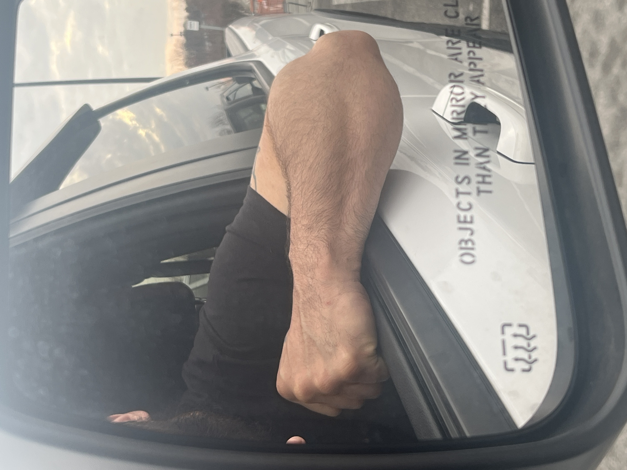 The image shows the view in a car's side mirror. The mirror reflects the driver's arm and hand, which is gripping the car door. The arm is hairy and muscular. The car door is white. The mirror also shows "OBJECTS IN MIRROR ARE CLOSER THAN THEY APPEAR." The background is blurred but appears to be a road or parking lot with a building and cloudy sky above. Based on the provided location, the image was taken in Orange Lake, United States.