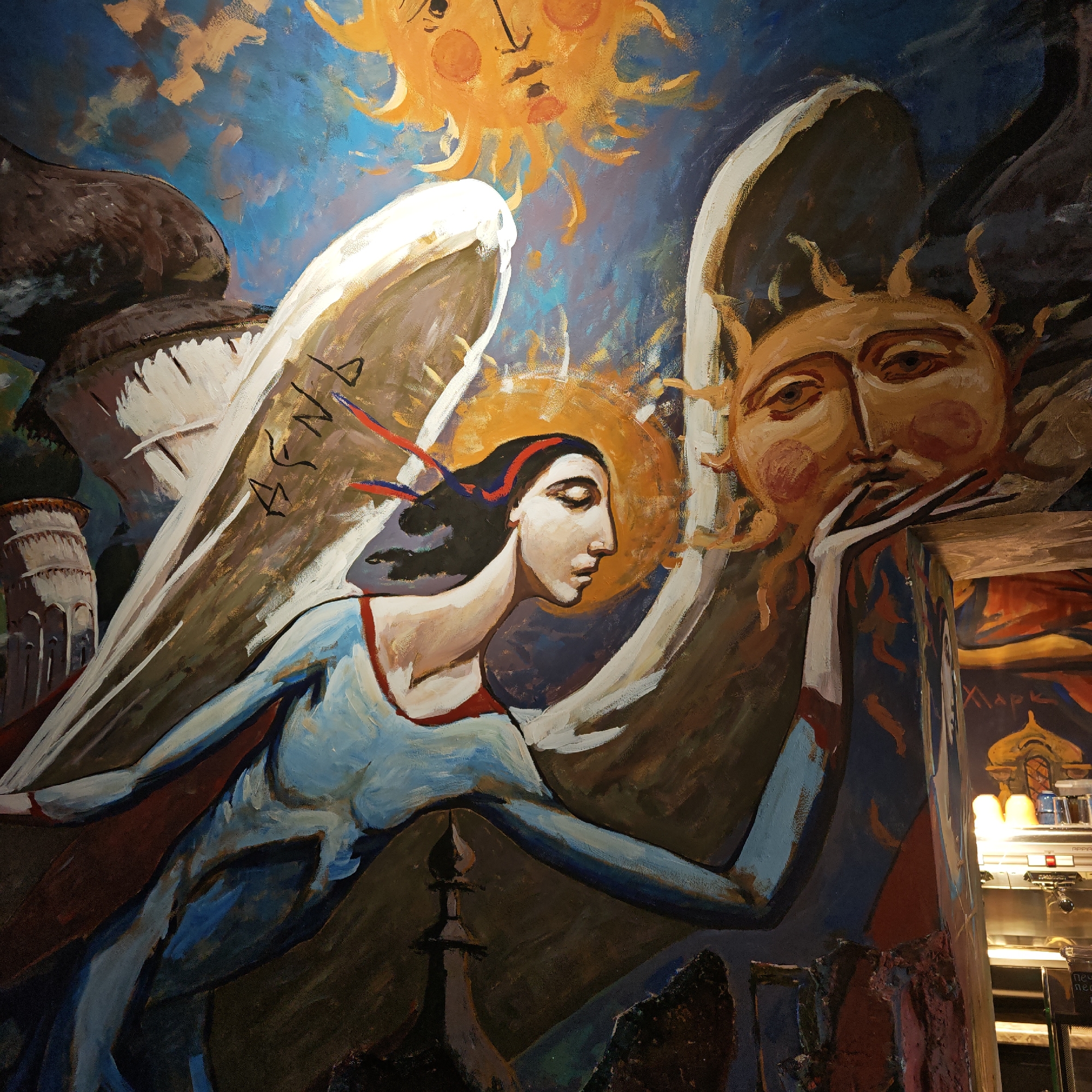 A painting on a wall depicts an angel-like figure with white wings and a halo behind her head. The figure has dark hair, a red headband, and is wearing a blue dress. She is positioned between two suns with faces. The painting also includes stylized architectural elements, including a tower resembling the Leaning Tower of Pisa. The wall appears to be part of a commercial space, as indicated by the presence of a coffee machine and other equipment visible in the background. The words "ХАРК" appear on the wall near the equipment.