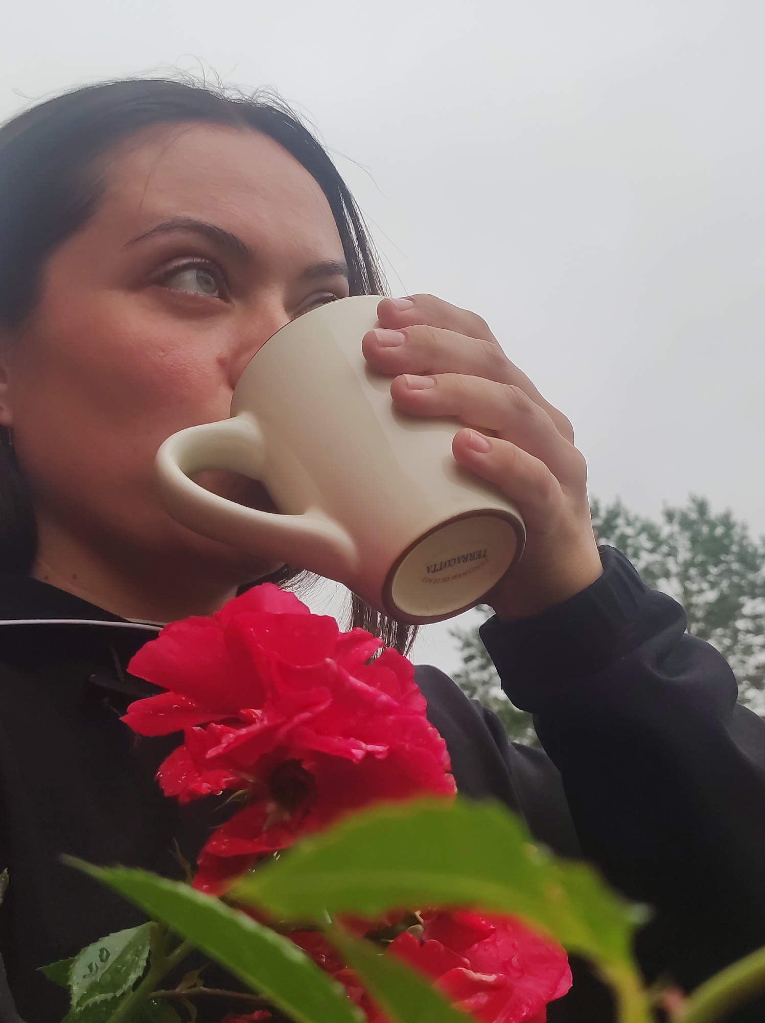 A medium shot captures a woman holding a white mug and drinking from it, with a bright red rose and green leaves in the foreground. The woman has dark hair and is looking to her left. She is wearing a black hooded sweatshirt. The background is blurred and appears to be an outdoor setting with trees visible and a cloudy sky. The text "TERRACOTTA" is visible on the bottom of the mug. The image does not contain any information that would allow for the identification of a specific location within the city of Фоки, Russia.