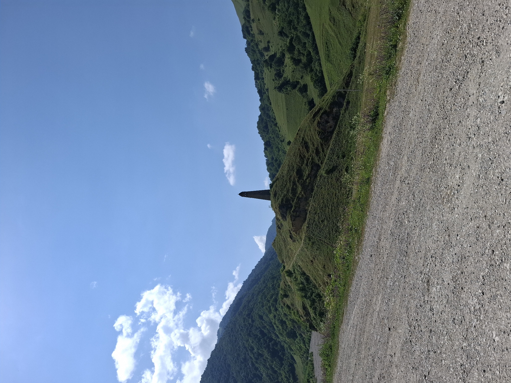 The image shows a tall, slender stone tower on top of a grassy hill. The tower is dark brown and appears to be made of rough stone. The hill is covered in green grass and some patches of exposed earth. A dirt road is in the foreground, with gravel and small stones scattered across it. In the background, there are rolling green hills covered in trees, and a large, lush green mountain to the left. The sky is a clear blue with a few white clouds. The scene appears to be a natural landscape with a historical monument, likely in a rural or mountainous area. The specific location in Kharachoy, Russia, is not visually confirmed by the image alone, but it is described as the location. There are no people or animals visible in the image.