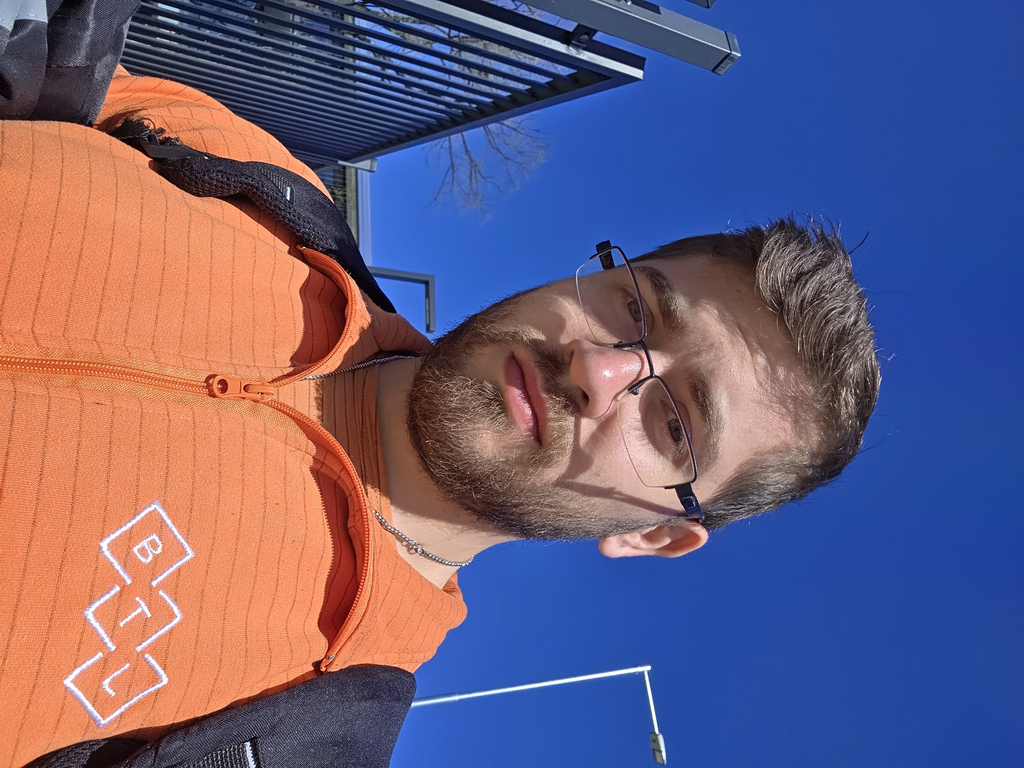 A man with a beard and glasses wears an orange jacket with the letters "BTJ" on it. He is facing the camera. The background shows a bright blue sky, a metal pergola, and bare tree branches. A lamppost is also visible in the lower part of the image. The location is Plovdiv, Bulgaria.