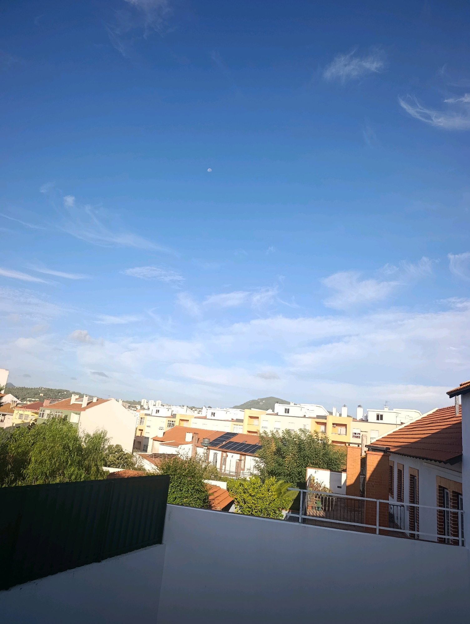 The image captures a clear blue sky with wispy clouds and a faint crescent moon visible in the upper portion. Below the sky, a cityscape of Setúbal, Portugal, is depicted, characterized by a dense cluster of residential buildings with terracotta roofs and white or light-colored facades. Trees with lush green foliage are interspersed among the buildings, suggesting a neighborhood with residential green spaces. The foreground is dominated by a white wall and a dark green fence, implying a private balcony or rooftop vantage point from which the photograph was taken. Solar panels are visible on the roof of one building, indicating modern infrastructure within the urban setting. The overall scene suggests a daytime view of a residential area in Setúbal, Portugal, under a bright sky.