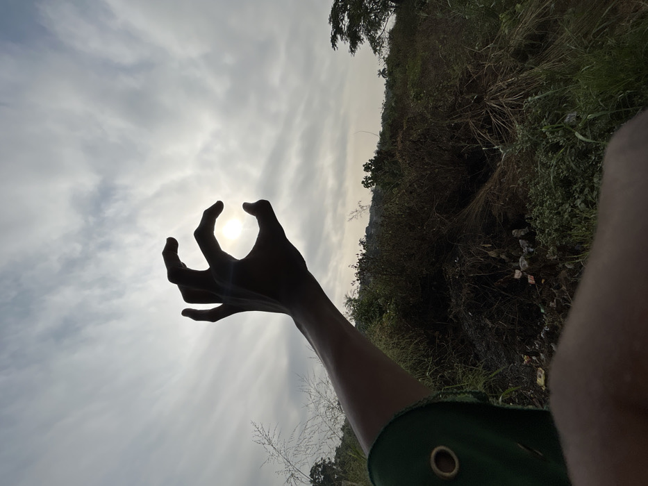 A vertically oriented outdoor photograph taken in the city of Debasco, Nigeria, features a human right arm and hand silhouetted against an overcast sky. The hand is positioned with the thumb, index, and middle fingers curved to frame the bright, visible sun, forming a "C" shape. The arm wears a green garment, with a brass-colored grommet visible on the sleeve near the wrist. The background consists of dense, dry vegetation, including tall grasses and shrubs, with scattered refuse present on the ground. Trees are visible in the mid-ground and distant background, suggesting a natural landscape or uncultivated area. The scene is heavily backlit by the sun.