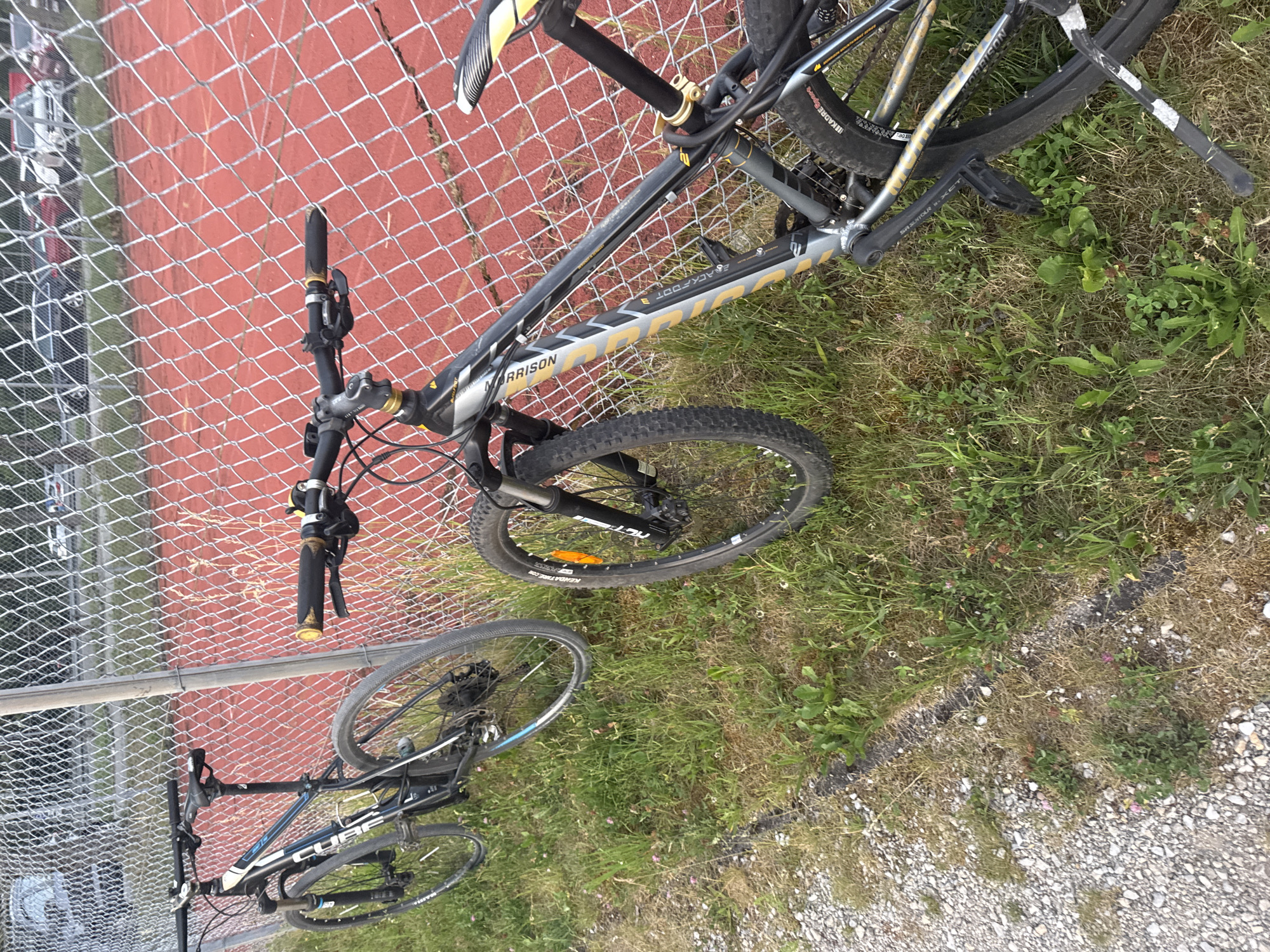 Two bicycles are parked next to a chain-link fence with a red surface visible behind it, likely a sports court. The bicycles are on a grassy area. The bicycle on the left is black and blue with "CUBE" written on the frame. The bicycle on the right is black and silver with "MORRISON" written on the frame. The background shows trees and vehicles, suggesting an outdoor public space. The scene is consistent with a location near the sports facilities of Neubiberg, Germany.