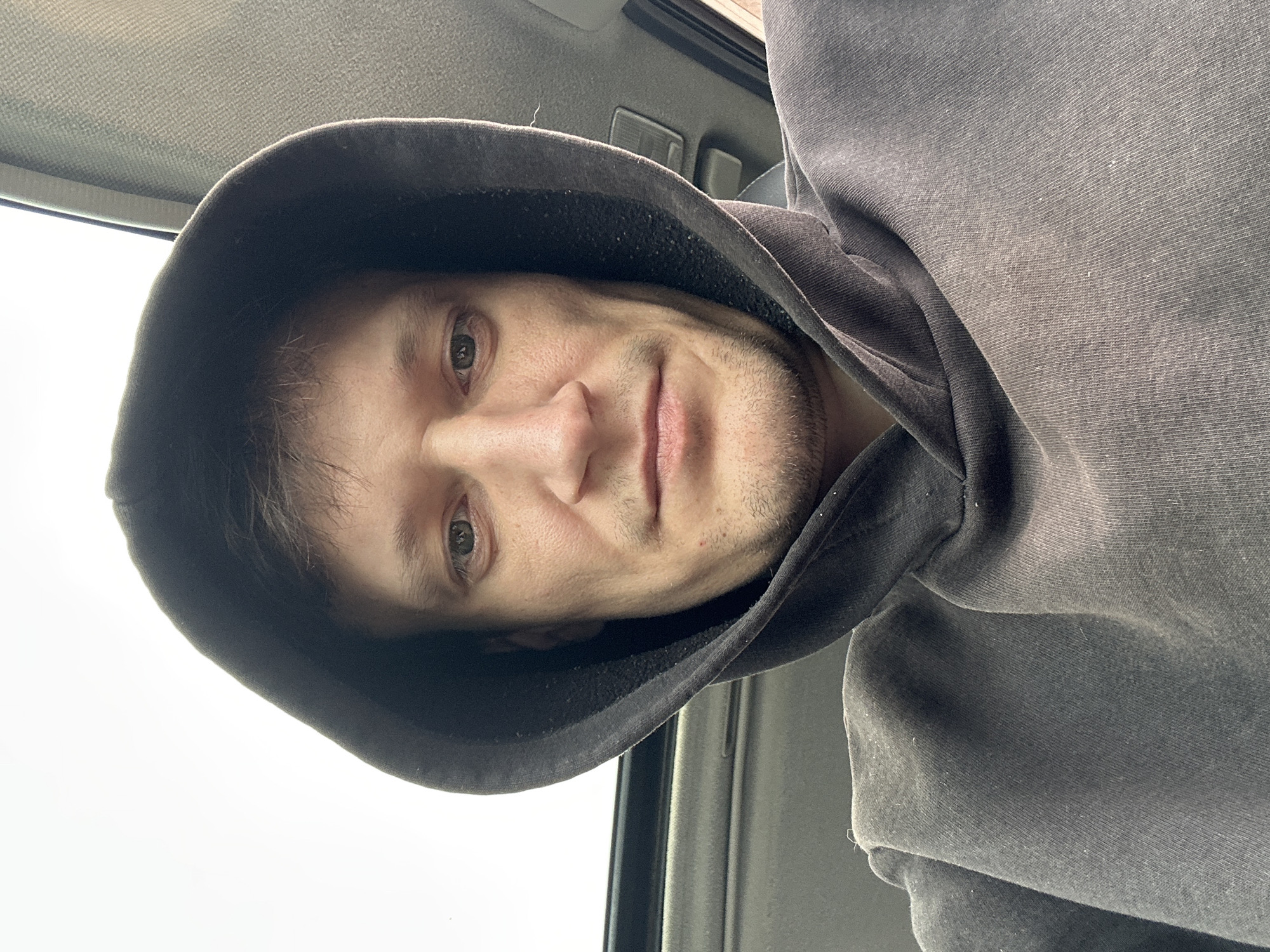 The image is a close-up, selfie-style shot of a man wearing a dark grey hooded sweatshirt. The man's face is centrally positioned and faces the camera. He has short, brown hair, fair skin, and light-colored eyes. He has stubble on his chin and jawline. His left eye is looking directly at the camera, while his right eye is slightly averted. The hood of the sweatshirt is pulled up, framing his face. The background appears to be the interior of a vehicle, with a sunroof visible at the top of the frame and what looks like car upholstery on the right side. The lighting suggests an overcast day. There are no discernible landmarks or features that would indicate the specific location within the city of Makiv, Ukraine.