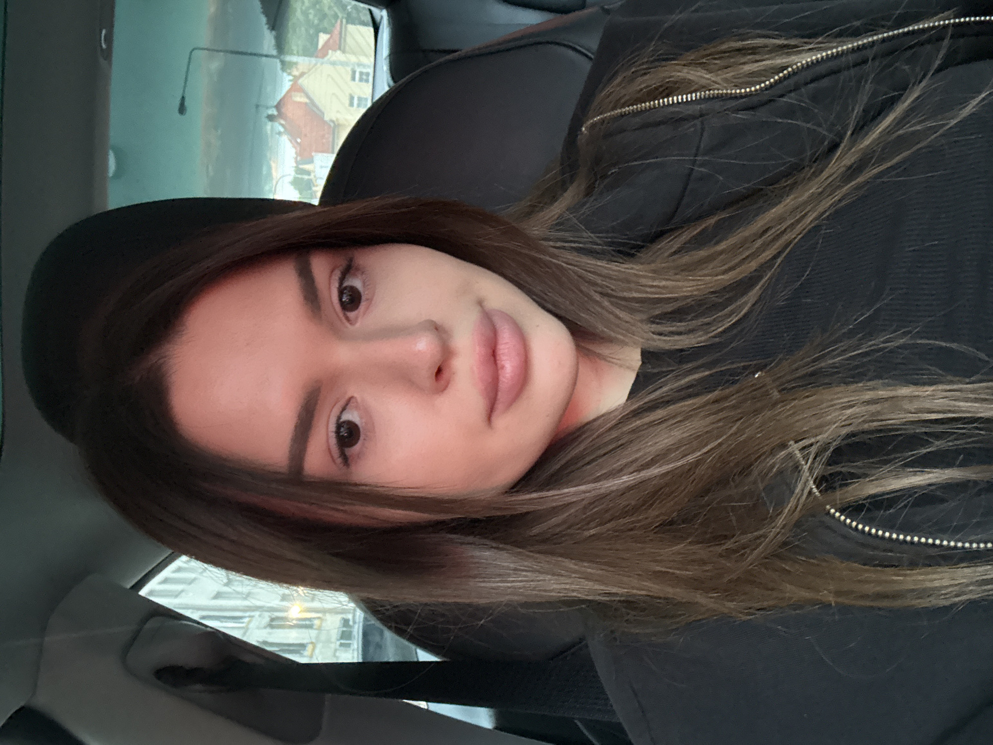 A young woman is taking a selfie in the front passenger seat of a car. She has long, dark brown hair and is wearing a black top and jacket. The background shows buildings and a streetlamp, suggesting an urban environment. The lighting indicates it is likely dusk or dawn.