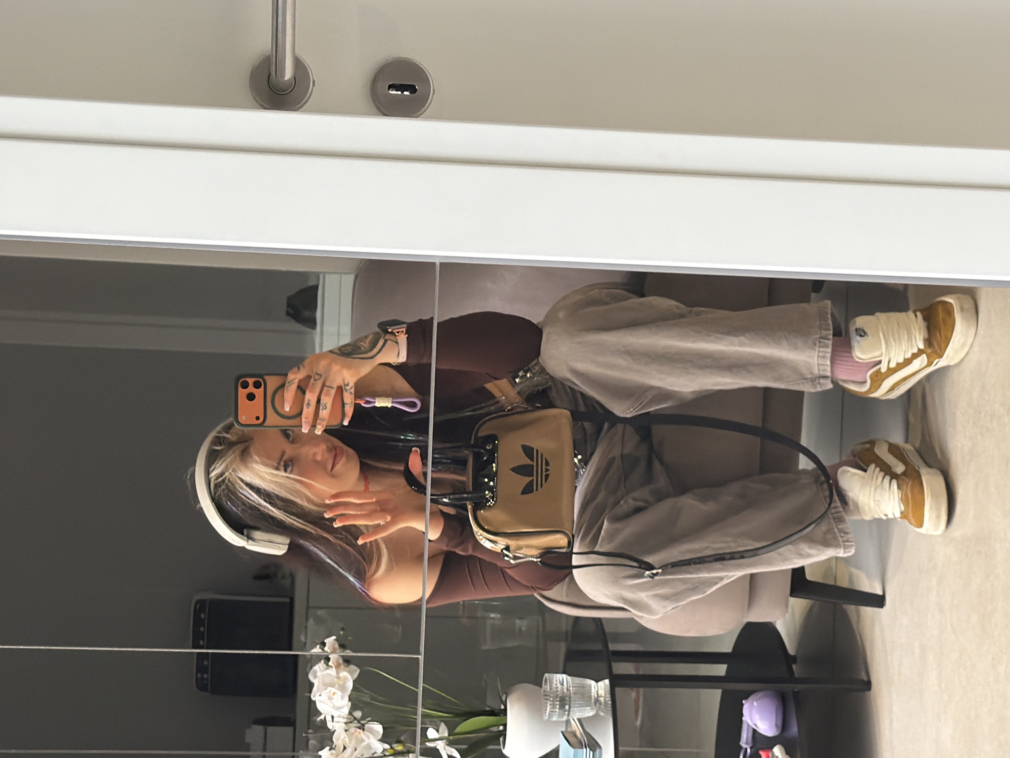 The image is a mirror selfie taken by a young person seated indoors in Budva, Montenegro. The subject is positioned centrally, looking towards the camera reflected in the mirror, and appears relaxed with a slight smile.
The person has long hair with dark roots and prominent blonde or light streaks framing their face. They are wearing large white over-ear headphones and a dark brown long-sleeved top paired with loose-fitting, light beige or greyish-brown pants. Their footwear consists of tan and white high-top sneakers, with light pink socks visible. On their left wrist, a watch is visible. Their left hand holds a smartphone with a distinct orange case, poised to take the picture, while their right hand is gently raised. Tattoos are visible on their fingers and arms.
A notable accessory is a small, rectangular Adidas handbag, tan in color with a black Adidas trefoil logo, worn across their body. The visible text on the bag is "adidas".
The background, reflected in the mirror, suggests a modern indoor setting. Above the person's head, a white door with silver door handles and a keyhole indicates a room entrance. To their left, a dark wall or surface is visible, along with a black speaker or electronic device. Part of a light-colored sofa or chair is seen on the right, where the person is seated. At the bottom right, a small table with black legs holds a vase containing white orchid flowers, a clear glass, and a small purple object. The scene conveys a casual yet stylish moment within a personal space.