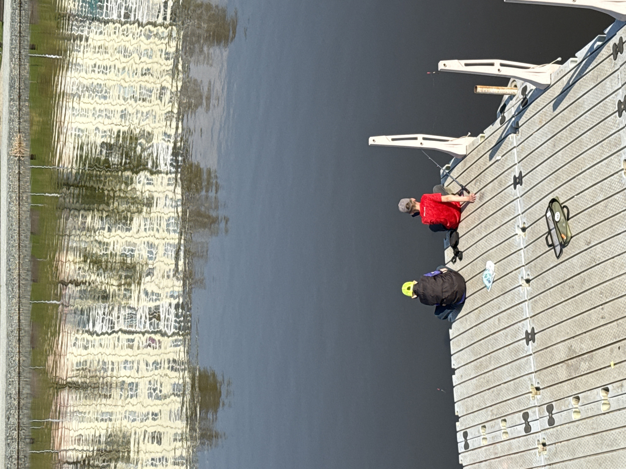 Two people are fishing from a floating dock on a body of water. One person wears a red shirt and the other a black jacket and yellow hat. Both are kneeling and facing the water. Fishing rods are placed in holders on the dock edge. A bag is near the person in the red shirt. The water reflects a building and trees. The location is in Votkinsk, Russia.