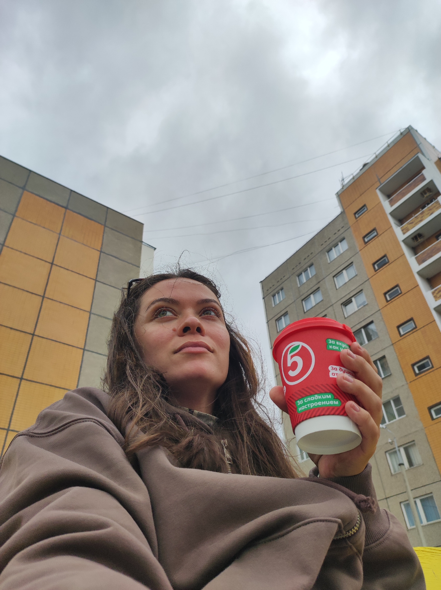In Chaikovsky, Russia, a young woman with long, wavy brown hair and green eyes is captured from a low angle, holding a red disposable coffee cup with a white lid. The cup has green and white text and a circular logo with a "5" inside a circle. She is wearing a brown hooded sweatshirt. The background features the corner of a modern apartment building with a yellow and grey facade, and another grey apartment building behind it. The sky is overcast with grey clouds, and power lines are visible crisscrossing the sky. The woman is looking upwards and to her left, with a thoughtful expression.