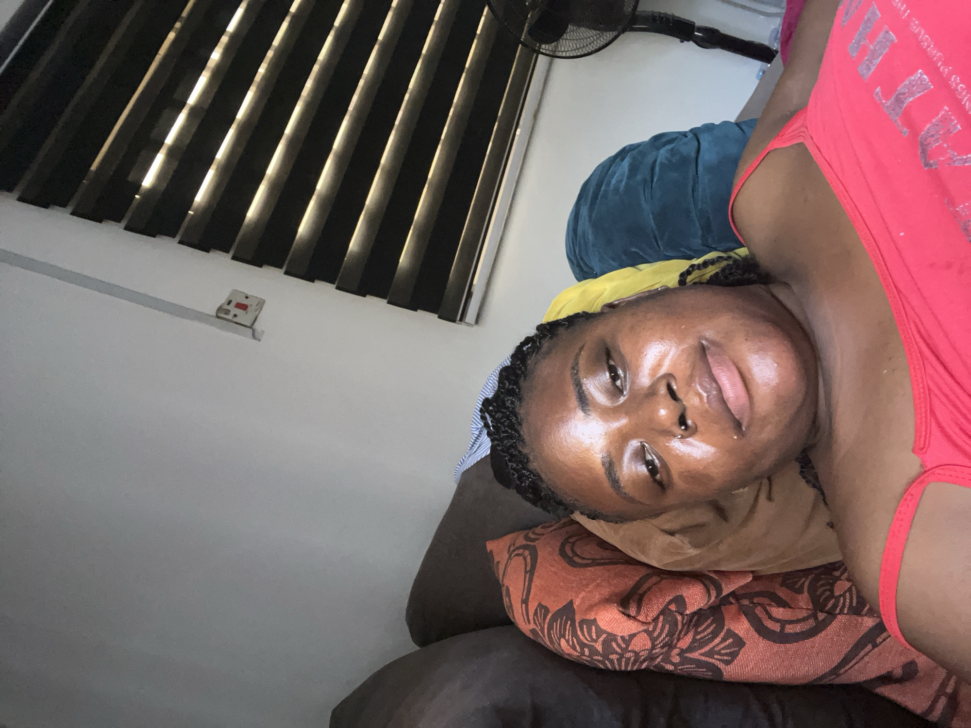 A young Black woman is lying on a couch in what appears to be an indoor setting. She is wearing a bright red tank top and has her hair braided. The couch is covered with pillows of various colors, including orange, yellow, and blue. In the background, there is a wall with a window covered by horizontal blinds. A light switch is visible on the wall. A black electric fan is positioned to the right of the frame. The lighting suggests it is daytime, with natural light filtering through the blinds. The overall impression is a casual, relaxed pose.