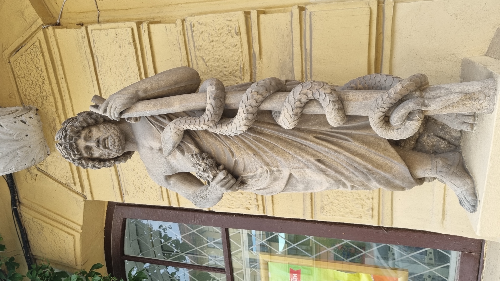 This is a medium shot, eye-level, outdoor photograph of a stone statue of a male figure with a snake coiled around a staff. The statue is located on the exterior of a building with a yellow facade. The statue depicts a muscular male with curly hair and a beard, wearing a draped garment. He is holding a bouquet of flowers in his left hand and grasping the staff with his right hand. The snake is wrapped around the staff, its head facing upwards. The statue stands on a stone pedestal. To the left of the statue is a dark-framed window with diamond-shaped panes. The building facade has horizontal decorative elements. The overall lighting suggests it is daytime. The statue is identified as Hygieia, a Greek goddess of health, and is located on the former Jewish hospital building in Chernivtsi, Ukraine.