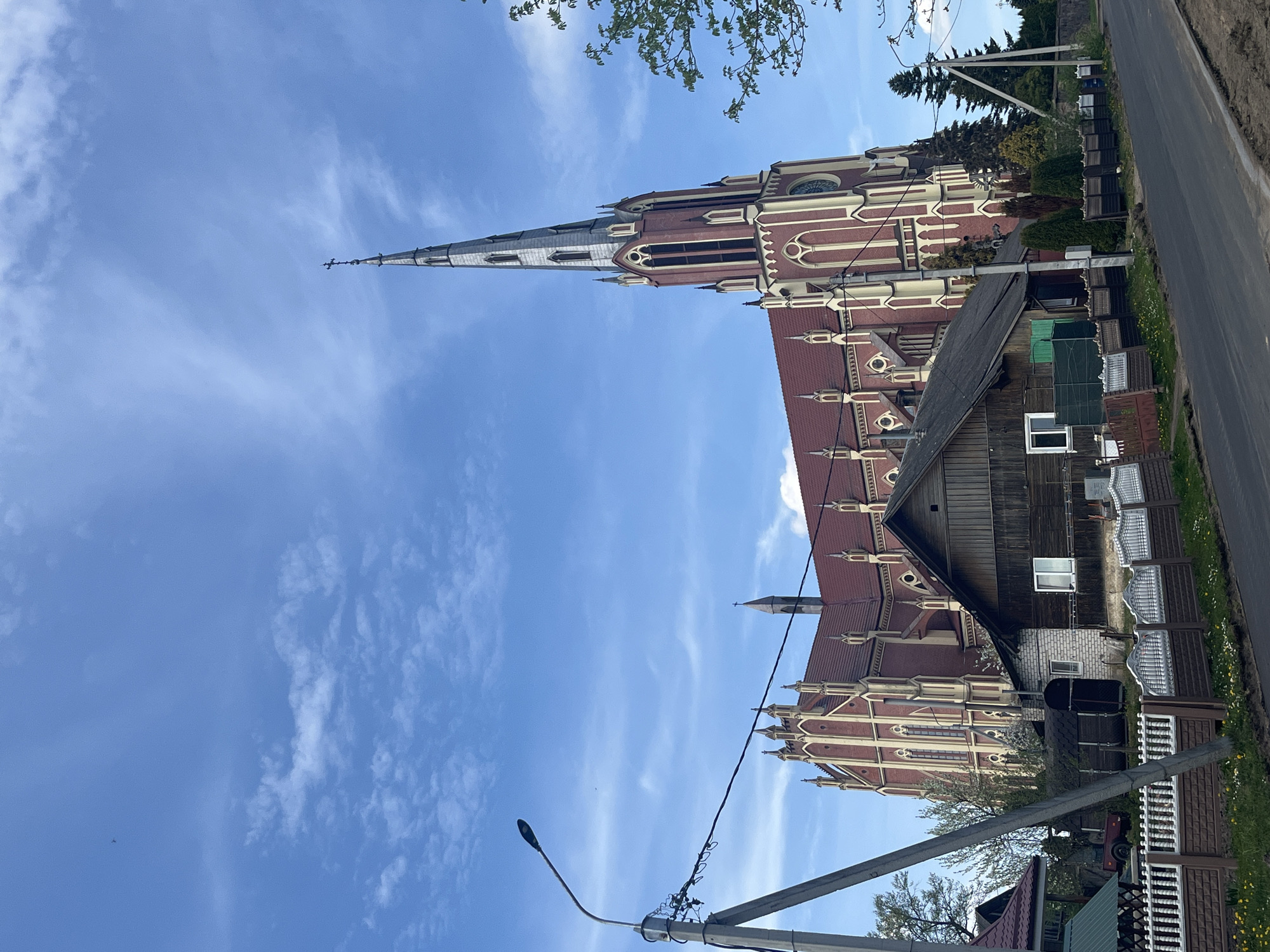 The image shows the Church of the Holy Trinity in Гервяцкі сельскі Савет, Belarus, captured from a low angle. The church, constructed of red brick with light-colored trim, features a tall, pointed steeple. Next to the church, there is a weathered wooden house with a dark roof. A road runs along the right side of the image. A utility pole with a street light and associated wires are in the foreground, partially obscuring the view. The sky is blue with scattered clouds. The surrounding area includes grass, fencing, and some trees.