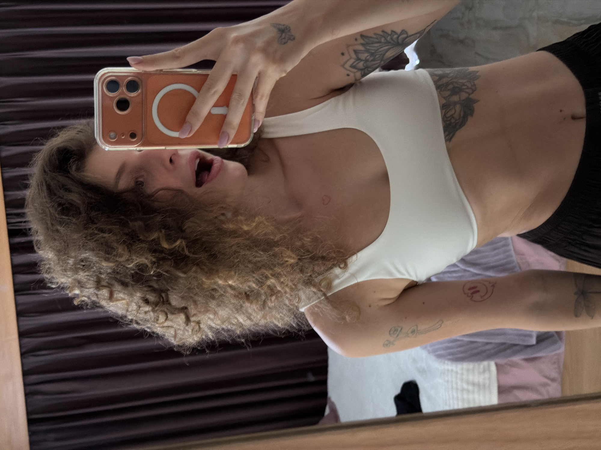 This sideways mirror selfie captures a young woman from her upper chest to her mid-thighs, taken indoors in North Kuta, Indonesia. She has voluminous, curly light brown hair and is in an expressive pose with her mouth open, suggesting surprise or playfulness. Her right hand, with light pink nail polish, holds an orange smartphone in a clear case, showcasing a three-lens camera system and a white circular MagSafe ring on the back.
She wears a white athletic-style crop top or sports bra and black ribbed shorts or pants. Several tattoos are visible: a small butterfly on her right wrist, a larger floral or mandala design on her right bicep, a tiny red heart outline on her neck, and a prominent floral tattoo on her right side below her breast. On her left arm, she has a small heart outline, a stick figure, a red cartoon-like symbol, and a dragonfly tattoo.
The background, reflected in the mirror, shows dark brown curtains or blinds on the left and various light-colored fabrics, including purple and white textiles, on the right. A thin wooden frame on the left indicates the mirror's edge. The ambient lighting suggests the photo was taken during the daytime. No visible text is present in the image.