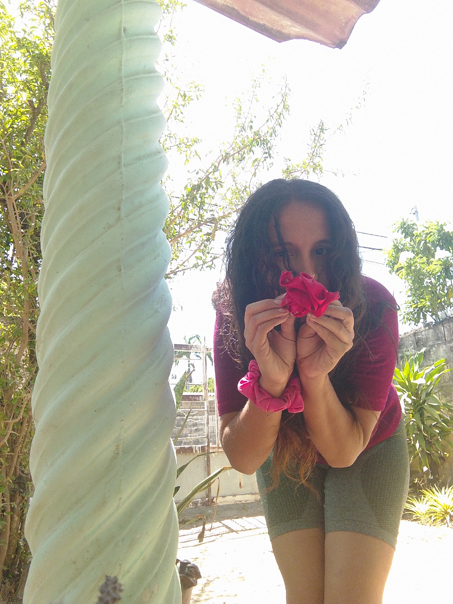 A woman with dark, curly hair is holding a red rose to her nose. She is wearing a maroon t-shirt and olive-green shorts. On her left wrist, she has a pink scrunchie. The woman is outside, next to a white, spiraled column. Behind her, there are various plants and trees, as well as a section of a concrete wall and what appears to be a metal fence or gate. The sky is bright, suggesting it is daytime. The location may be Juan Griego, Venezuela.