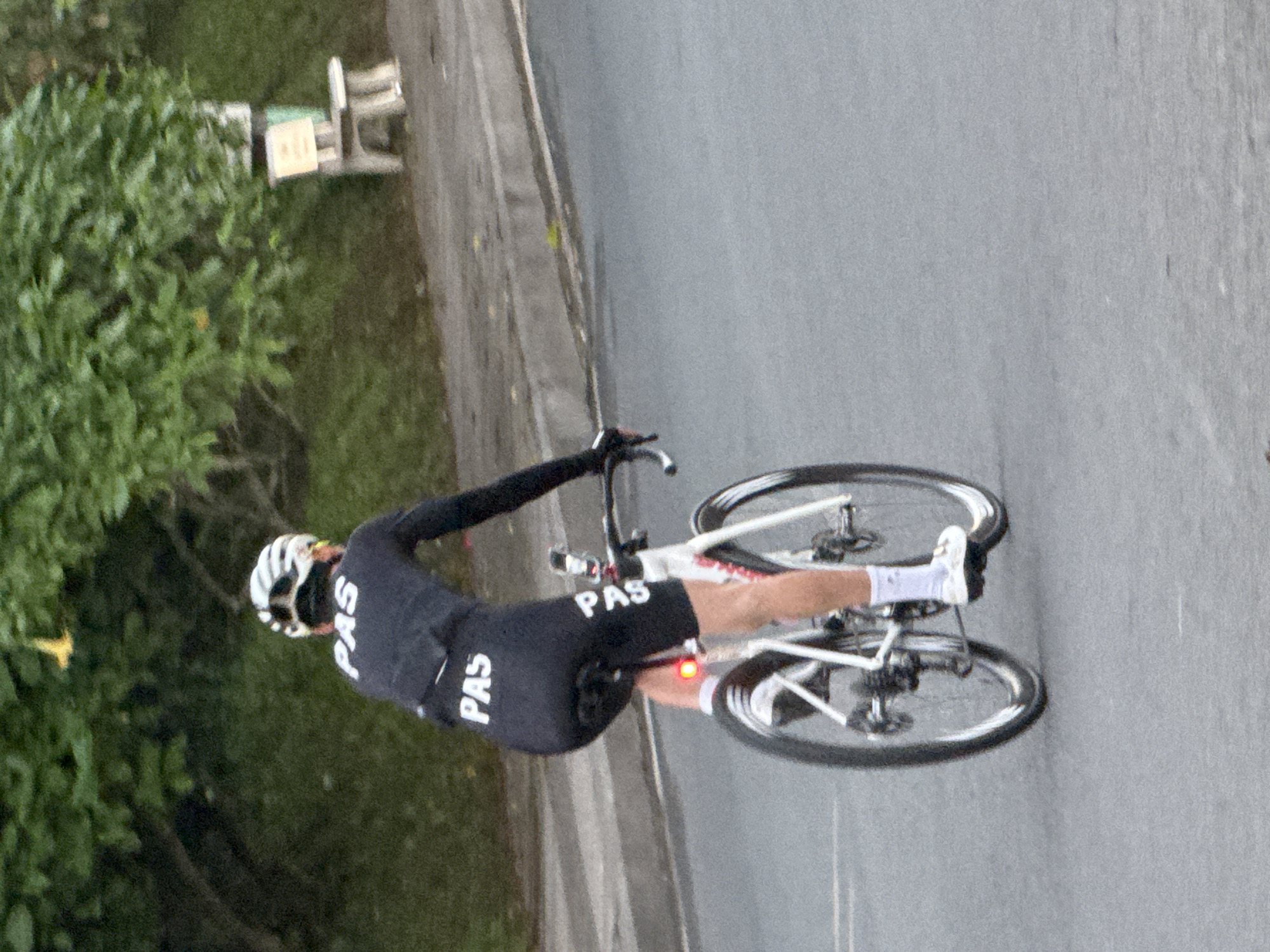 A cyclist, seen from behind, rides a white bicycle on an asphalt road. The cyclist wears a black cycling kit with "PAS" printed in white on the back of the jersey and on the left leg of the shorts. A white helmet with black accents is worn. The cyclist's left leg is visible, wearing white socks and white cycling shoes. A red taillight is attached to the bicycle's seat post. In the background, lush green foliage hangs over a grassy embankment. To the right, a light-colored wooden bench with a backrest is positioned near a grey post. The scene appears to be in Da Lat, Vietnam, based on the natural environment and the style of the bench.