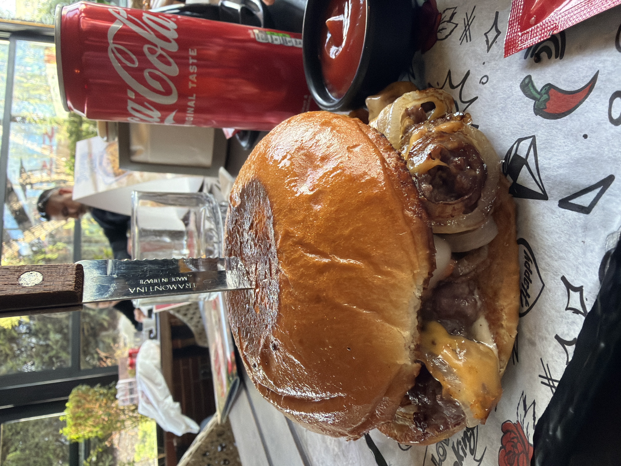 The image shows a partially eaten burger on a patterned paper, pierced with a knife labeled "Tramontina, Made in Brazil". A can of Coca-Cola and a small bowl of ketchup are nearby. The scene is indoors, possibly a restaurant, with a blurred view of an outdoor area visible through a window. A person is seen in the background, seated near the window. The location is identifiable as the city of Varna, Bulgaria, based on the context.