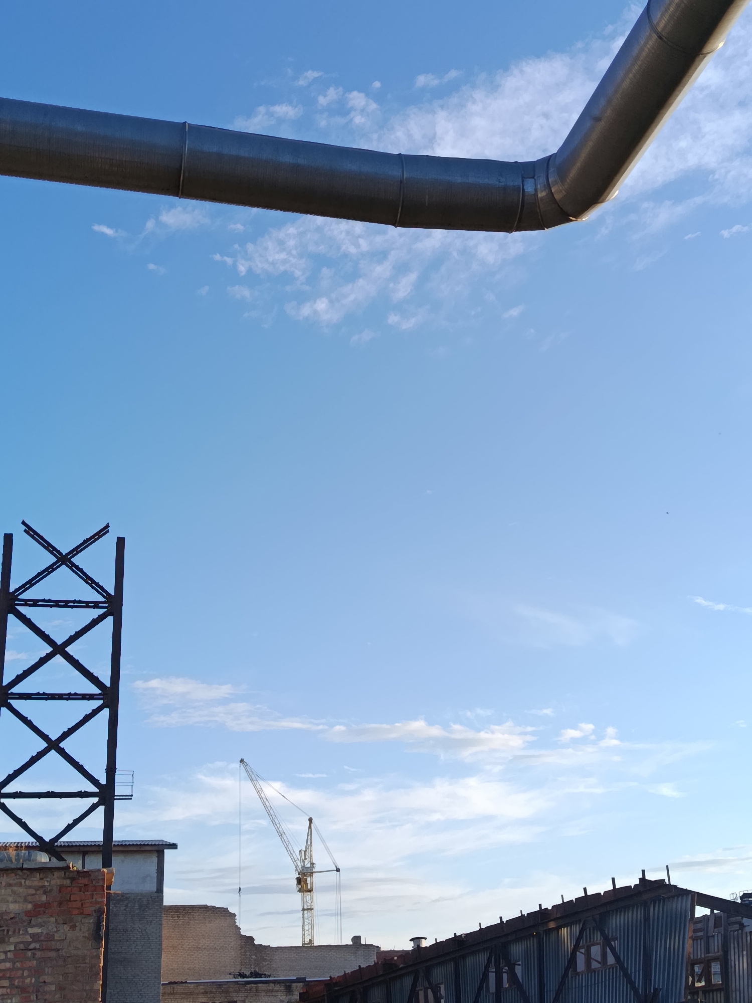The image depicts an outdoor scene with a clear blue sky and scattered white clouds. In the foreground, a large, metallic pipe extends across the upper portion of the frame. Below this pipe, a dark, angular metal structure, resembling scaffolding or a tower, stands to the left. Further to the right, a yellow construction crane is visible in the distance, positioned above several low-rise buildings. The buildings appear to be constructed of brick and corrugated metal, with some showing signs of disrepair. The scene suggests an industrial or construction area, possibly in the city of Svetlogorsk, Belarus, given the context provided. No people are clearly visible in the image.