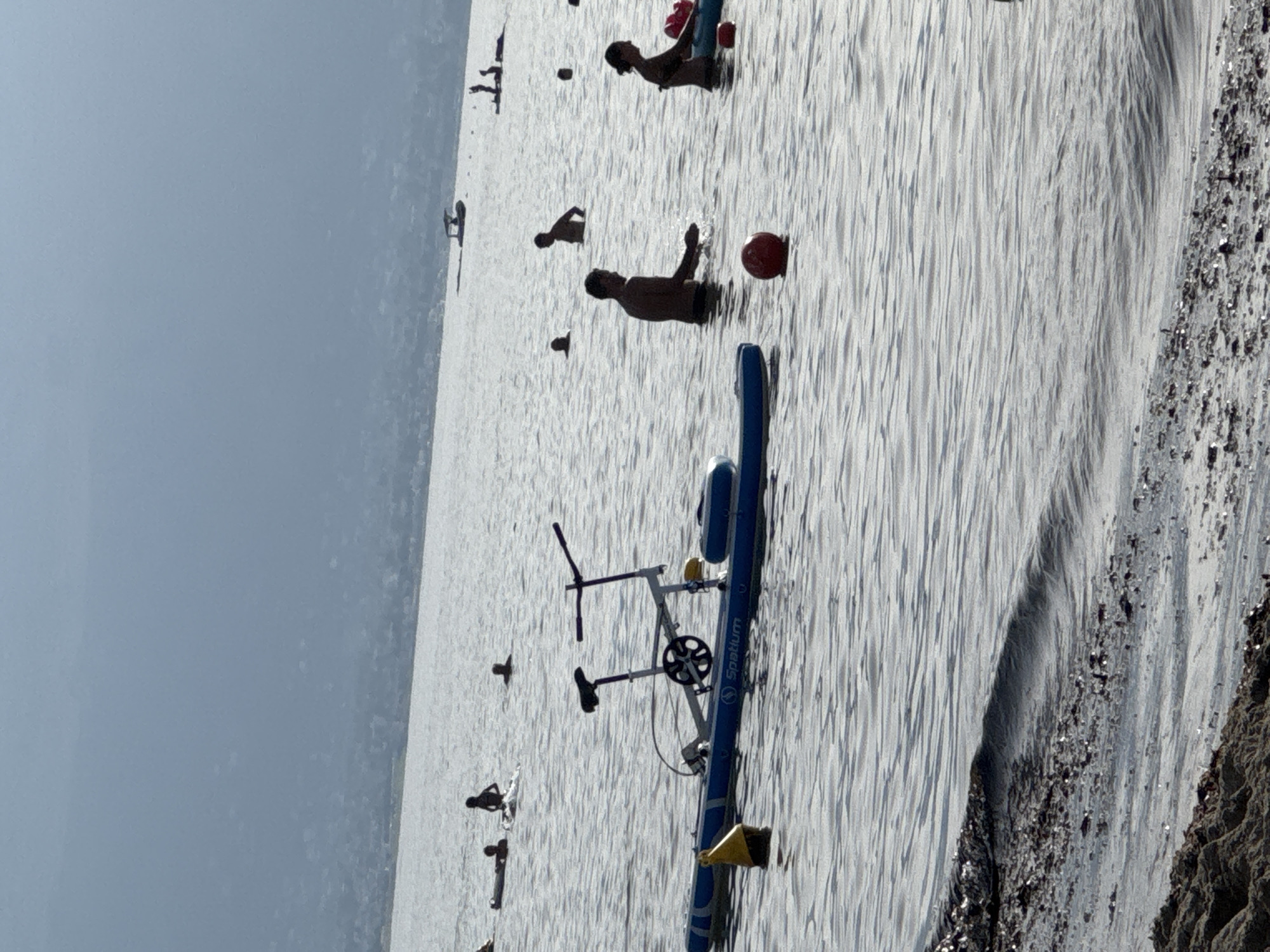In Isola delle Femmine, Italy, an inflatable watercraft with a bicycle mechanism is positioned on the water's surface. Several individuals are visible in the water, some partially submerged. The scene is set against a hazy background depicting a distant shoreline with buildings and a body of water. The foreground of the image shows the edge of a beach with dark sand and debris.