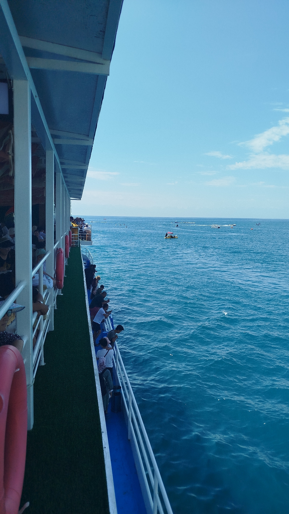 The image shows a view from the deck of a large white boat, looking out at a vast expanse of blue ocean under a clear sky with some scattered white clouds. The boat's white railing runs along the right side of the frame, separating the deck from the water. Several people are visible on the deck, leaning over the railing and looking out at the sea. The deck itself has a green carpet-like surface and blue painted sections. Life preserver rings are attached to the side of the boat. In the distance, several smaller boats and people on jet skis are scattered across the water, suggesting a recreational area or tourist destination. The overall impression is one of a bright, sunny day at sea, with passengers enjoying a boat ride.
