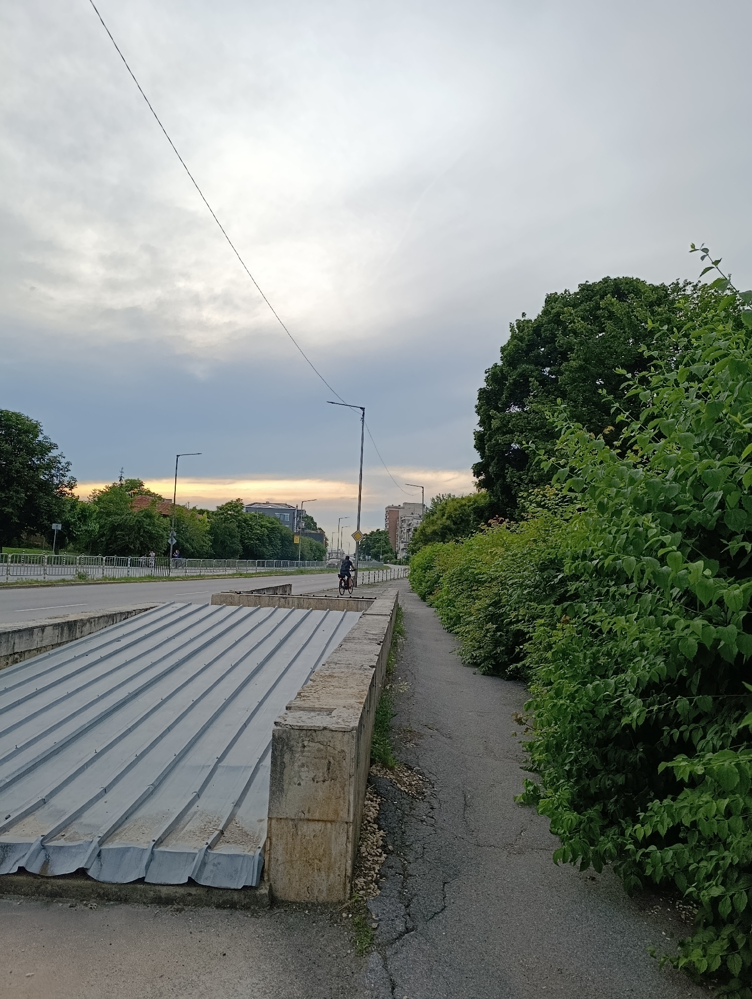 The image depicts a street scene in Razgrad, Bulgaria. A paved road runs horizontally across the frame, with a sidewalk on the right side bordered by lush green foliage. A person is seen cycling on the road. In the foreground, there is a gray corrugated metal structure, possibly a cover for an underground passage or infrastructure. The sky is overcast with hints of sunset colors in the distance. Buildings and trees are visible along the road.