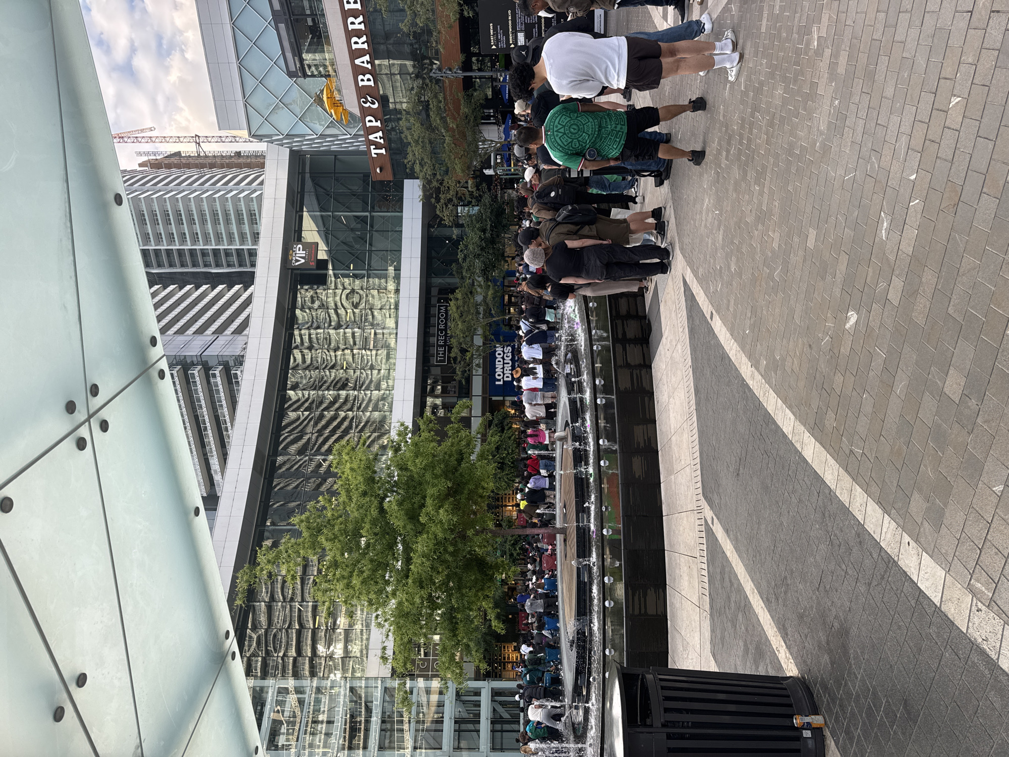 In Burnaby, Canada, an outdoor scene depicts a plaza with a fountain in the foreground and a large crowd of people gathered around it. The plaza is paved with brickwork and features a modern architectural design with glass-fronted buildings and a translucent overhead canopy. A mature tree stands in the center of the fountain area, surrounded by a circular wooden platform.
The crowd appears to be queuing or congregating, with individuals of various ages and backgrounds. Some are standing, while others are seated on the ground or on portable chairs. The people are dressed in casual attire, suggesting a public event or gathering.
Behind the fountain and crowd, the background reveals a bustling urban environment. Tall, modern buildings with reflective glass facades rise into the sky. Signage visible on the buildings includes "VIP," "THE REC ROOM," "LONDON DRUGS," and "TAP & BARREL," indicating commercial establishments. Construction cranes are also visible in the distance, suggesting ongoing development.
The overall atmosphere of the scene is one of a lively public space with a significant number of people engaged in an activity centered around the fountain. The weather appears to be overcast or cloudy.