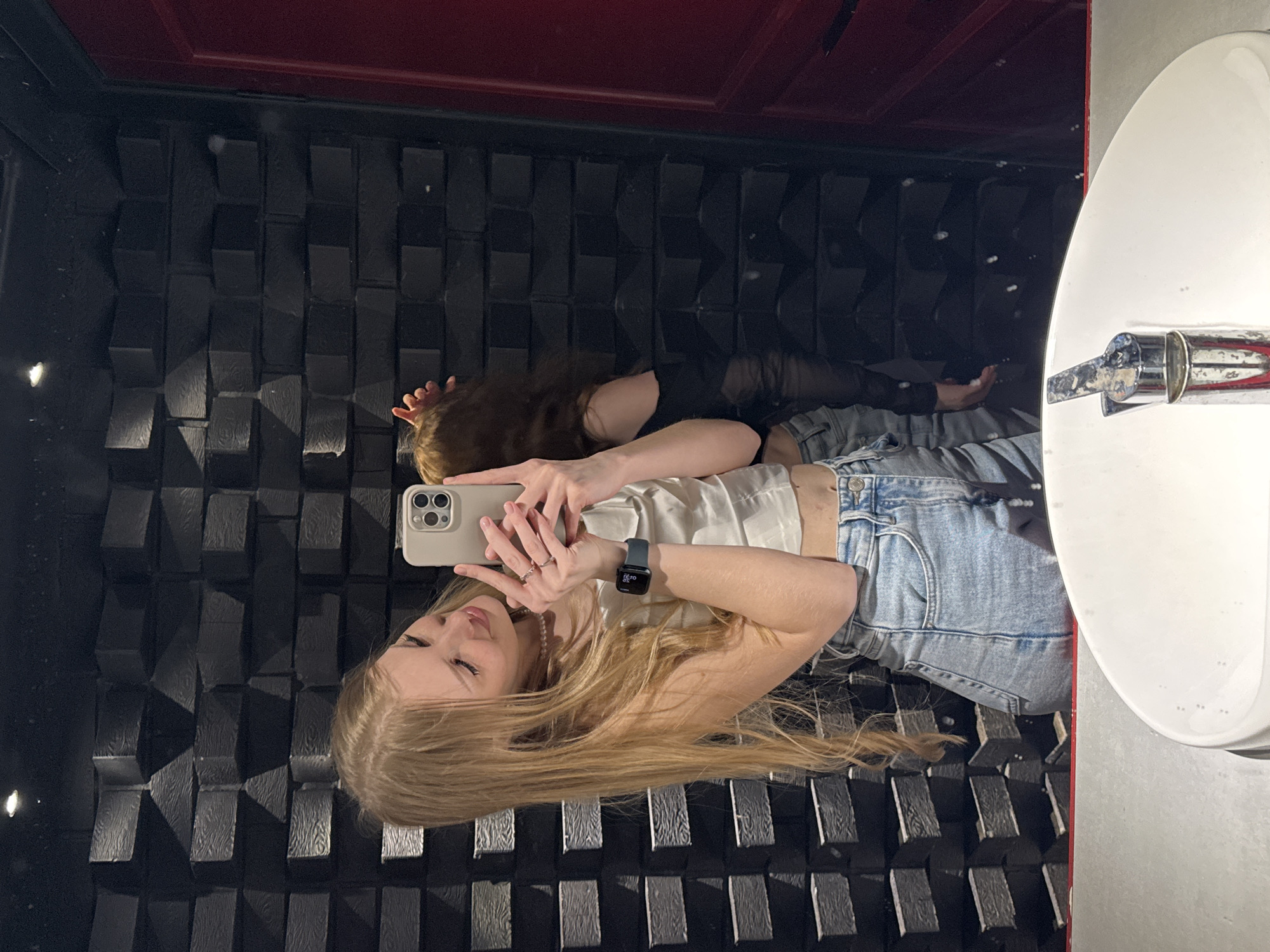 In a dimly lit restroom, a young woman with long blonde hair is taking a mirror selfie. She is wearing a white crop top, light blue jeans, and a smart watch on her left wrist. She is also wearing rings on her fingers and a pearl necklace. Her right hand is holding a smartphone, and her left hand is partially visible near her face. In the reflection, another person with long brown hair and wearing a black off-the-shoulder top is visible behind her, with their hands raised. The restroom features a textured black brick wall and a red door. A white sink with a chrome faucet is in the foreground. The image does not contain information that would allow for the determination of its location in the city of Perm, Russia.