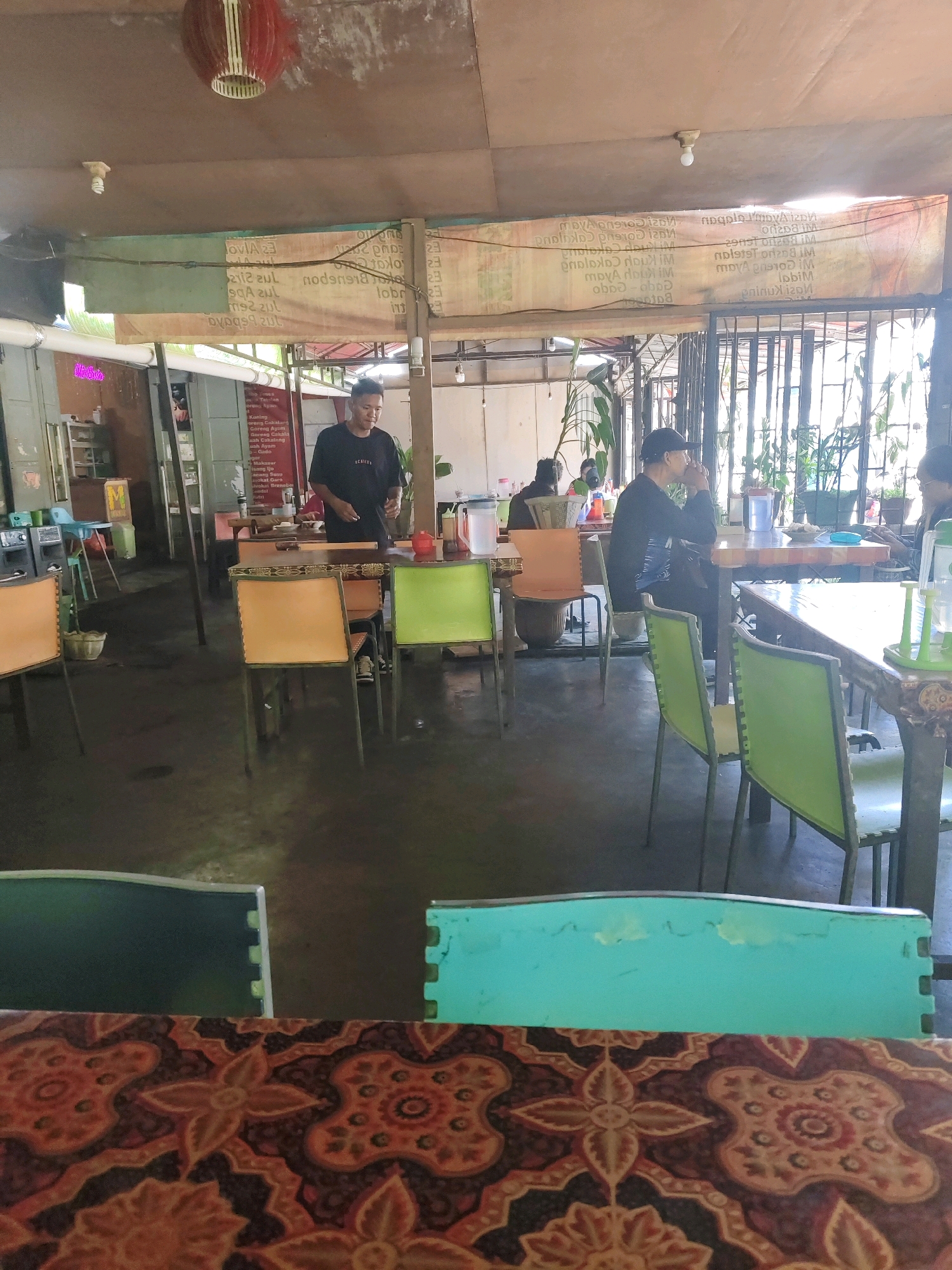 The image depicts the interior of a restaurant, possibly located in Tomohon, Indonesia, characterized by its open-air setup with a corrugated roof. A man in a black t-shirt is standing behind a counter, appearing to serve food or drinks. Several tables with chairs are visible, some occupied by patrons, suggesting a dining atmosphere. The environment is casual, with a mix of indoor and outdoor elements. Plants are present, adding a touch of greenery to the space. The overall impression is that of a local eatery or warung, potentially serving Indonesian cuisine. The flooring appears to be concrete or similar material. The lighting is a combination of natural light coming from the open sides and artificial lighting from fixtures on the ceiling. The image seems to capture a moment in time during the operational hours of the restaurant.