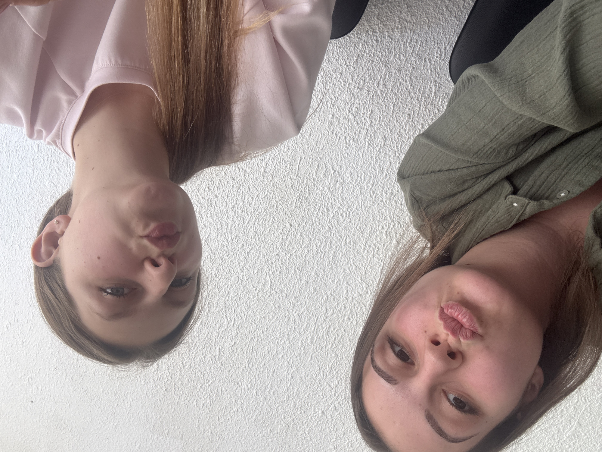 Two young women are posing for a selfie against a textured white wall. The woman on the left, closer to the camera, has brown hair and is wearing a green collared shirt. She is puckering her lips. The woman on the right, further from the camera, also has brown hair and is wearing a light pink shirt. She is also puckering her lips. Both women are looking at the camera. The scene appears to be indoors, possibly an office or common area, given the presence of black chairs visible in the foreground. There is no indication of the city of Burgas, Bulgaria, in the image itself.