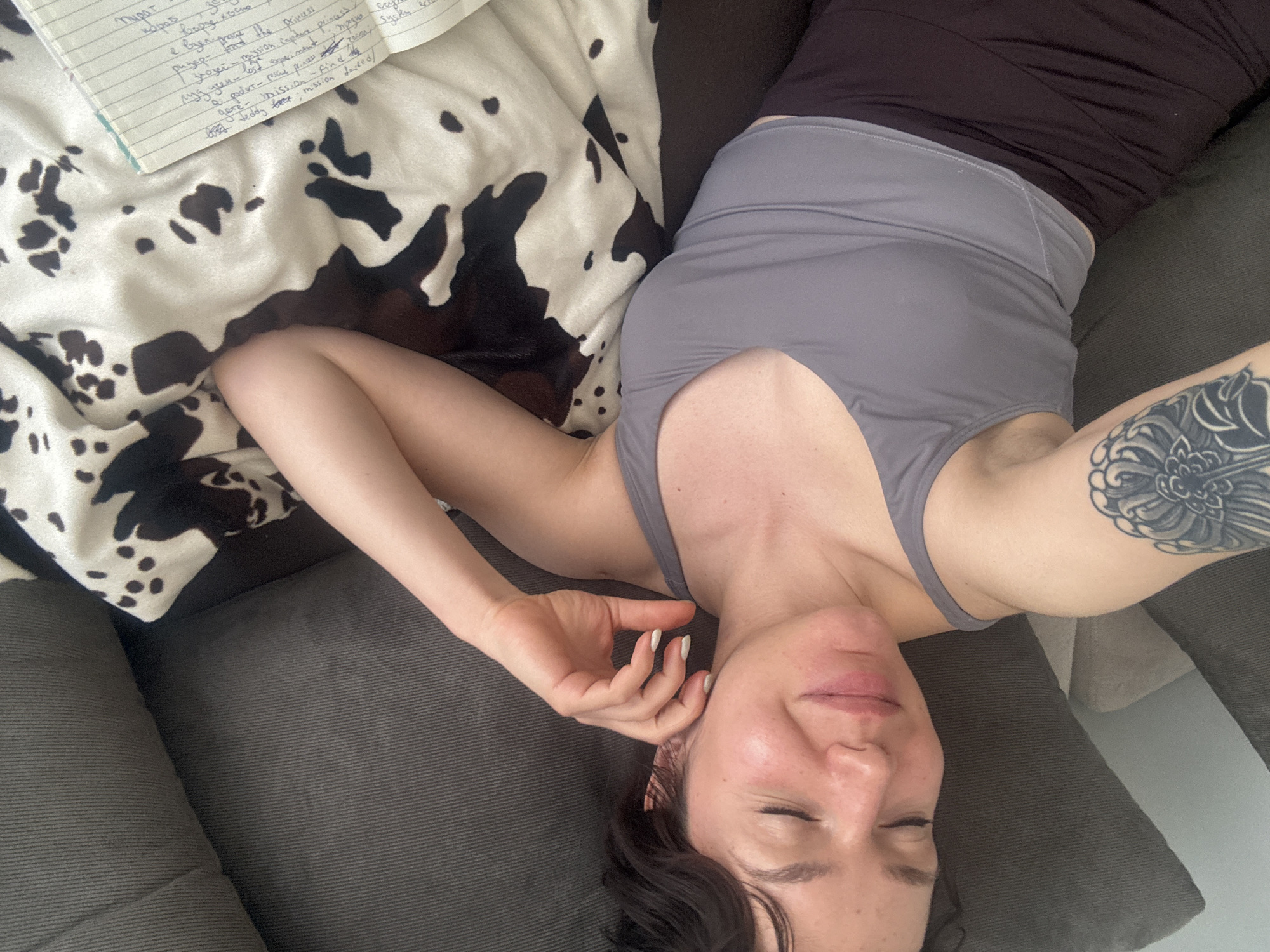 A woman with closed eyes is lying on a gray couch. She is wearing a black tank top under a gray cropped tank. She has a tattoo on her right upper arm. To her left is a cow-print blanket and a notebook. The text in the notebook is written in Russian. The photo appears to be taken from an overhead perspective. No location in Burgas, Bulgaria can be ascertained from the image.