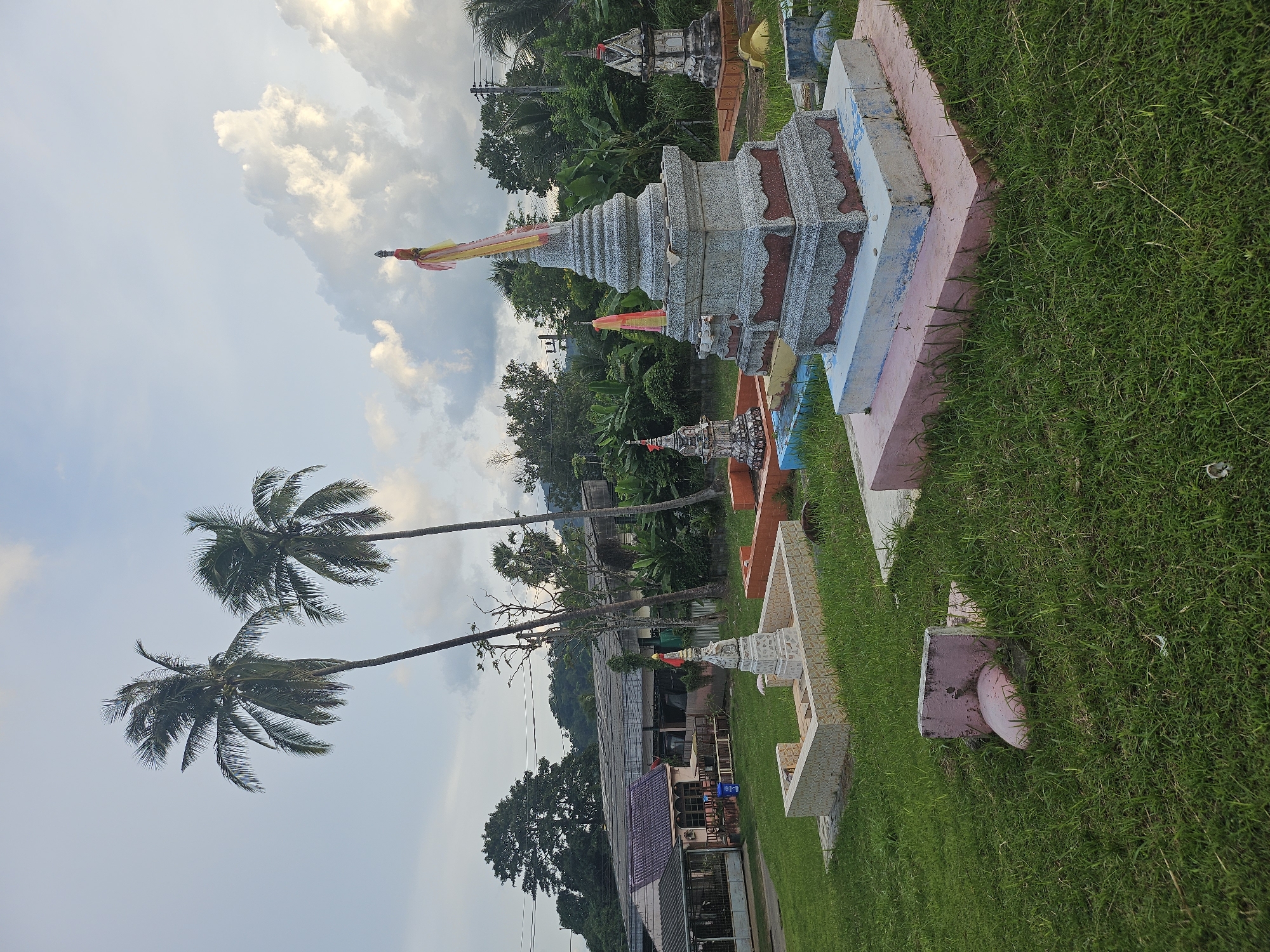 This media file depicts a row of decorative, tiered structures in an outdoor setting, likely a memorial garden or cemetery. The structures are made of stone and concrete, with some adorned with colorful fabric streamers, possibly signifying religious or cultural practices. They are arranged in a line, with a well-maintained grassy area bordering them on the right. In the background, lush green tropical vegetation, including palm trees, suggests a warm climate. Beyond the greenery, several buildings with tiled roofs are visible, indicating a nearby village or town. The sky is partly cloudy, with patches of bright sunlight breaking through, suggesting it might be late afternoon or early evening. The overall mood of the scene appears serene and contemplative. The image is oriented vertically, with the sky and palm trees at the bottom and the memorial structures at the top. No people are visible in the frame.