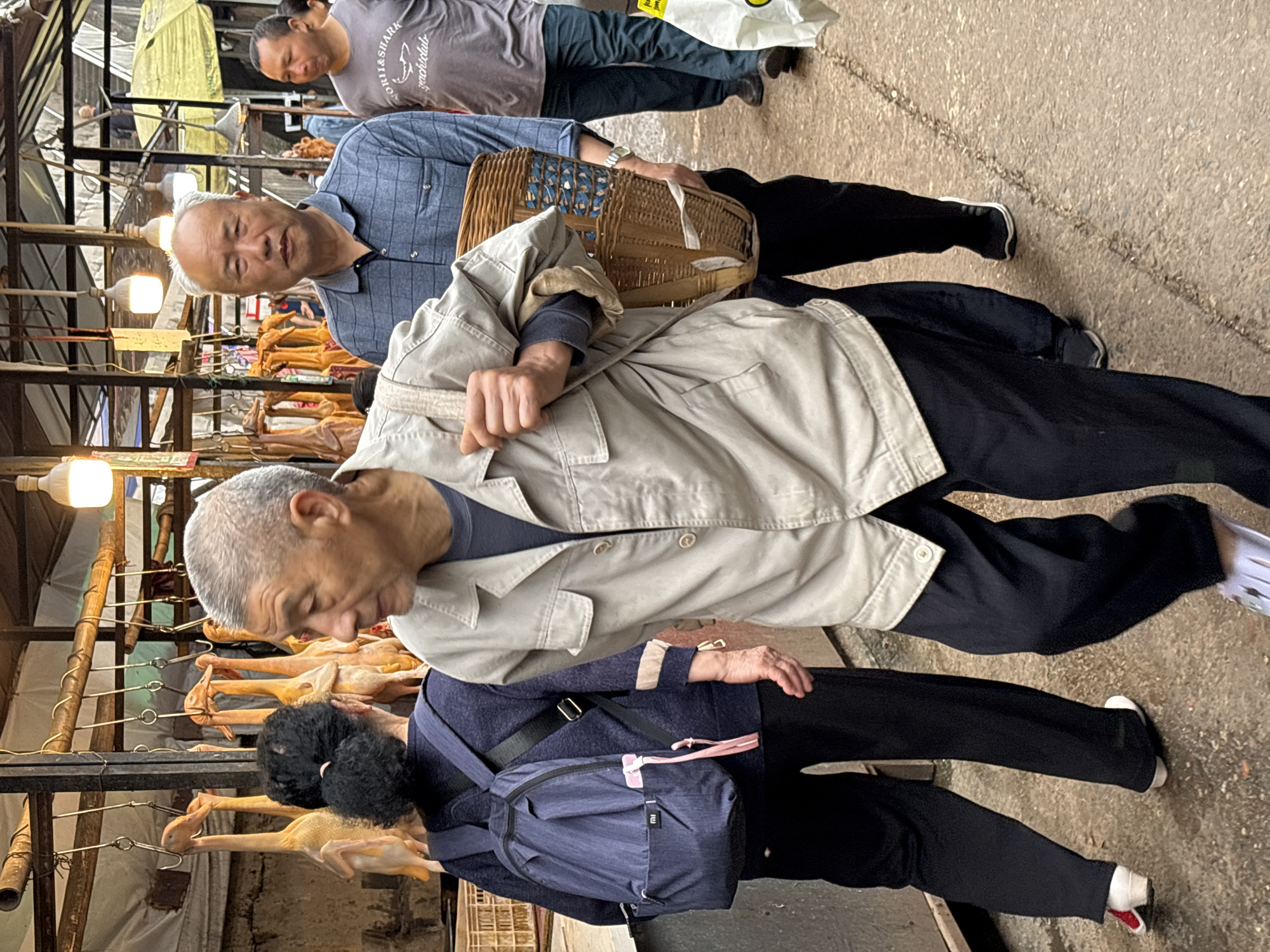 In Jiangjin, China, an outdoor market scene is captured. Dried poultry is displayed on metal rods under hanging light bulbs. An elderly man with gray hair, wearing a gray jacket over a blue shirt and black pants, examines the poultry. Behind him, an elderly woman with a black backpack also looks at the display. To the right and above these two individuals, another man, wearing a blue checkered shirt and carrying a woven basket, also views the poultry. At the top of the image, a man in a gray shirt is also visible. The flooring is concrete. The scene gives the impression of a traditional, outdoor market setting.