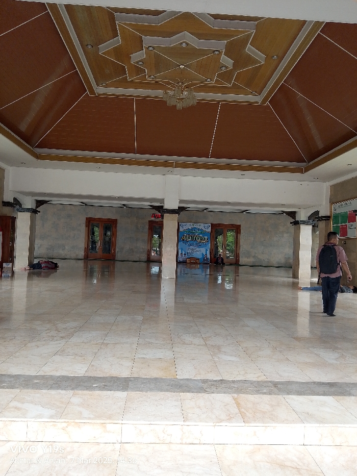 The image shows the interior of a building with a white tiled floor and light brown walls. The ceiling is covered in brown wooden panels, forming a star-shaped pattern with a chandelier in the center. There are large windows and doors on both sides, letting in natural light. There are several people in the room, including a man with a backpack walking towards the camera. The room is empty, with the exception of the people and a few pieces of furniture. This is likely a mosque or prayer room. The location is in the city of Indramayu, Indonesia, at the coordinates -6.327 latitude and 108.322 longitude.