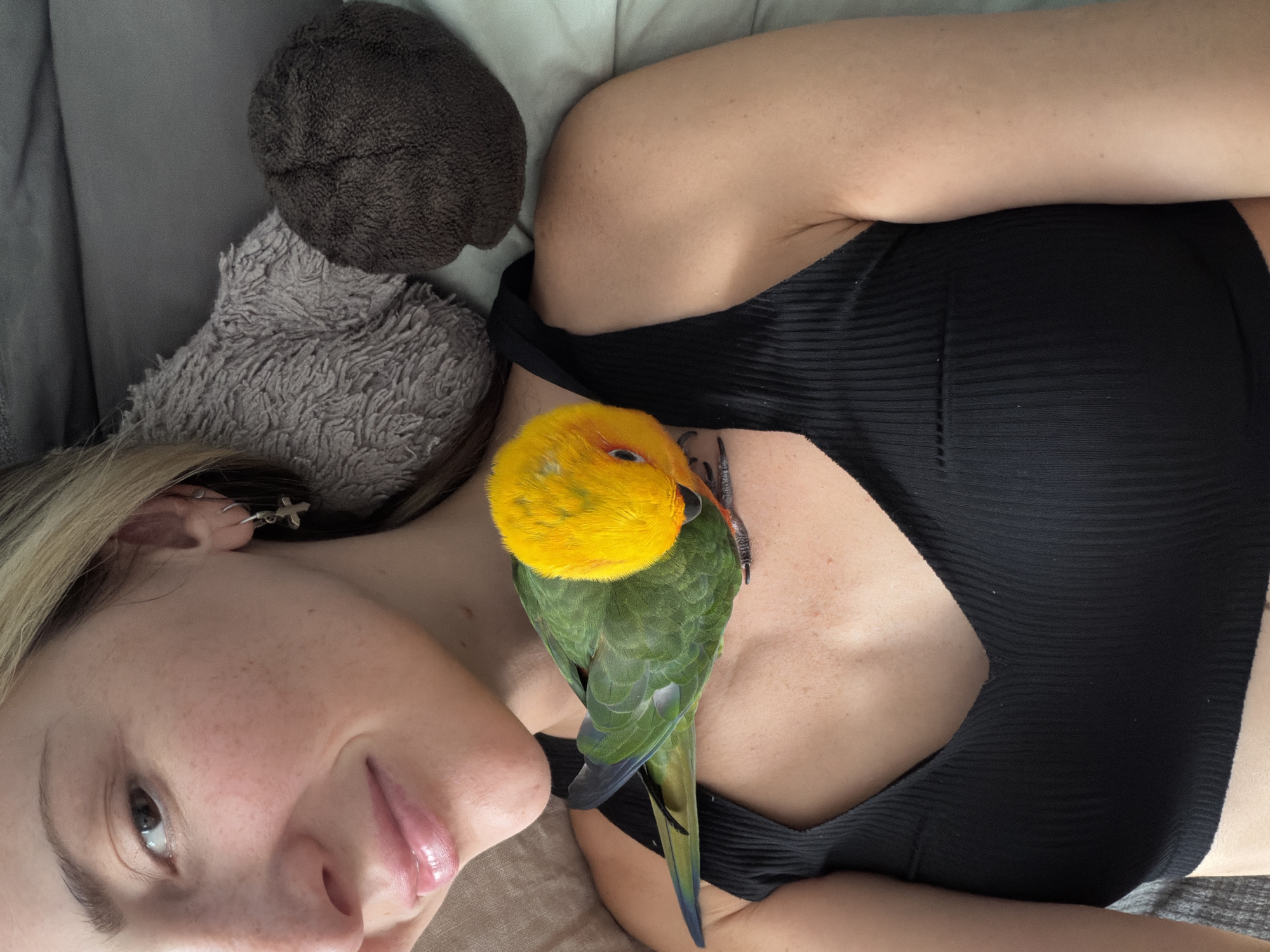 A woman lies on a surface with her head to the left of the frame. She has fair skin, blonde hair, and wears a small silver cross earring. She is wearing a black tank top. A colorful bird with a yellow head and green body perches on her chest. A gray, fuzzy pillow and a dark brown object are to the upper left of her head. The location in Khimki, Russia is not evident from this image.