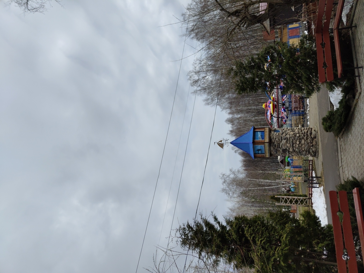 The image shows a park scene in Unknown. The sky is overcast. A decorative tower with a blue roof stands in the middle of the park, surrounded by trees. A carousel and benches are also visible. The ground has some snow patches. Power lines stretch across the sky.