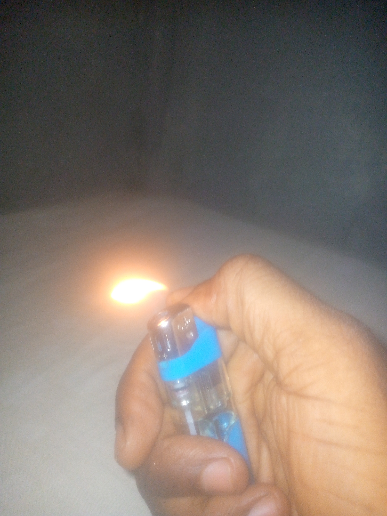 A person is holding a blue and clear lighter, which is lit, with their right hand. The background is blurry and indistinct, but appears to be a gray wall over a pale surface. The location is unknown.