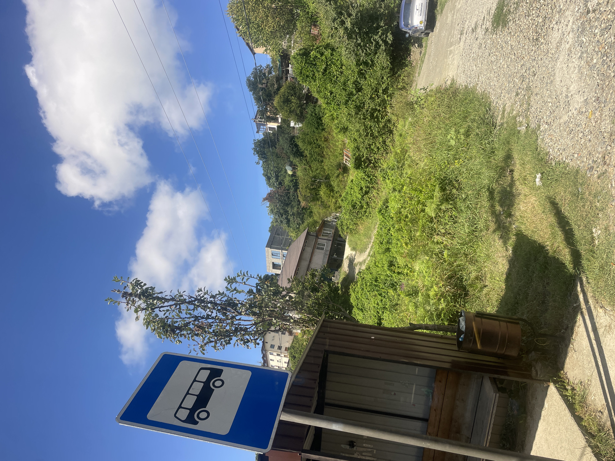 A bus stop sign is visible on the left side of the frame, with a bus icon on a blue background. Adjacent to the sign is a simple bus shelter with a wooden bench. A trash can is located next to the shelter. The shelter is situated on a concrete pad with grass and a gravel path leading away from it. The path winds up a steep, densely vegetated hill. Several houses are scattered among the trees on the hillside. A light-colored car is parked on the gravel path in the upper right portion of the frame. The sky is clear blue with scattered white clouds. The overall scene depicts a rural bus stop in Baranovka, Russia.