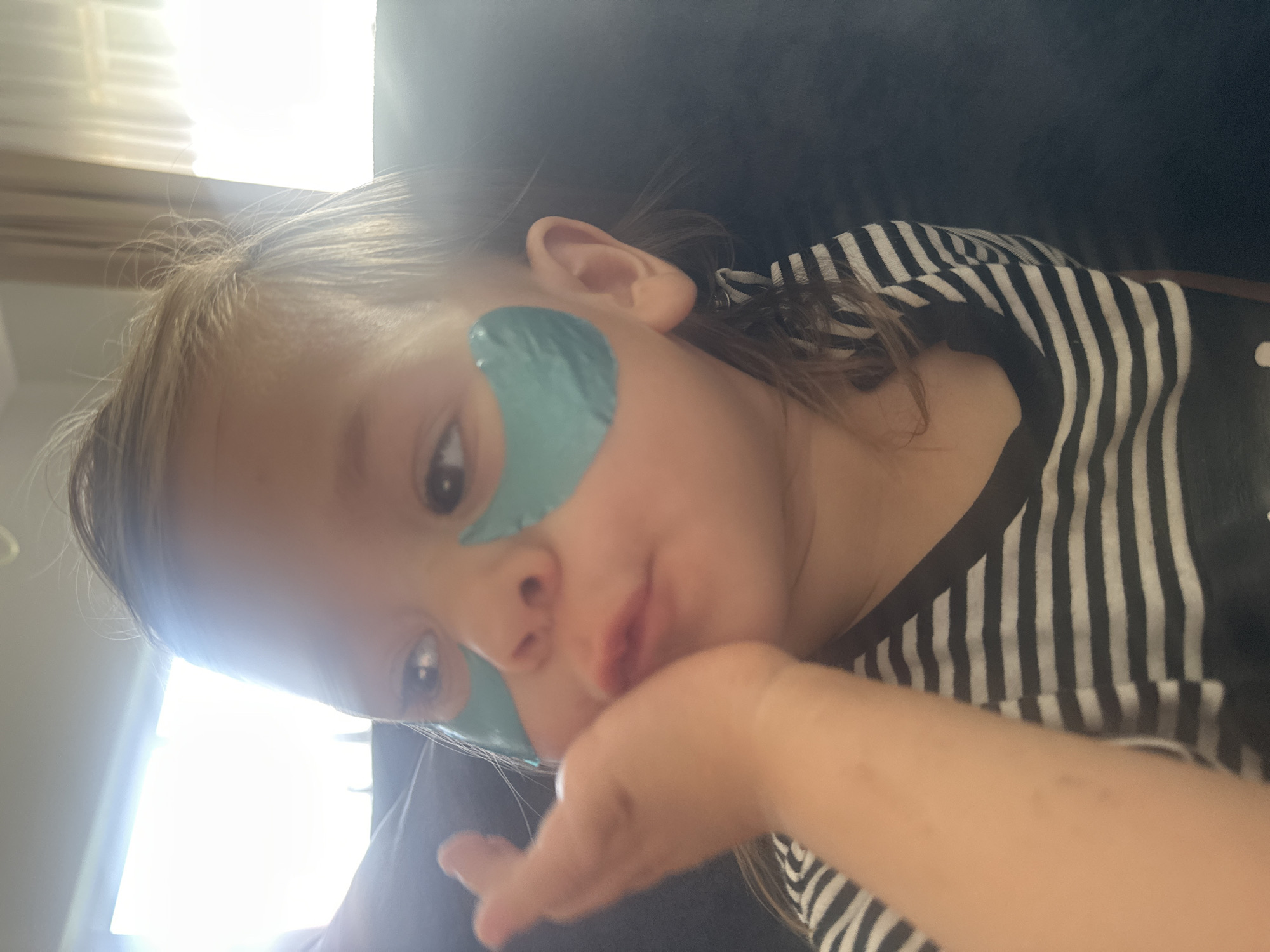 A young child, possibly a toddler, is seen in a close-up, profile view, looking to the left of the frame. The child has blonde hair and is wearing a black and white striped shirt. There are two teal-colored under-eye patches on the child's face. The child's left hand is raised to their cheek. The background is out of focus and appears to be an indoor setting with soft lighting coming from what looks like a window or sheer curtains. No specific location in the city of Kharnavy, Russia can be determined from the image.