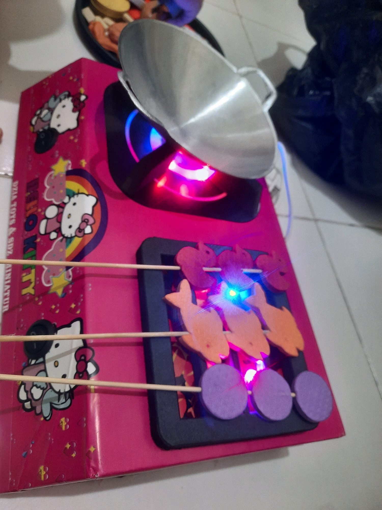 In a close-up, ground-level shot of a toy kitchen appliance in North Jakarta, Indonesia, the pink body of a play stove features Hello Kitty graphics. A small, silver toy wok rests on top, positioned above a lit-up area with pink and blue LEDs. In front of the stove, three toy skewers, each with colorful, fish-shaped pieces and purple circular pieces, are arranged. The background shows a blurred black object and a white tiled floor.