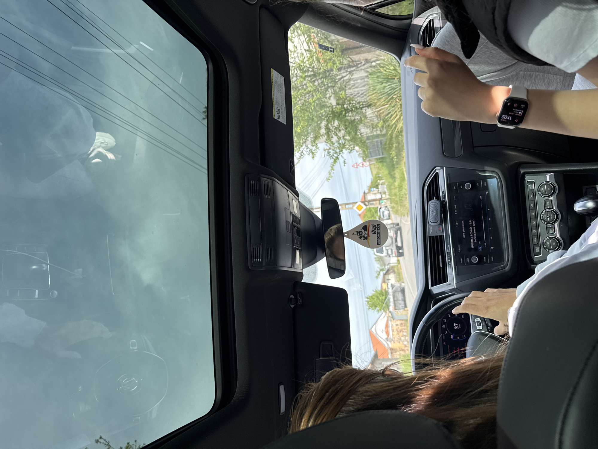 The image shows an interior view from the back seat of a car, looking forward. The car is in motion, with a speedometer reading of 37. The driver's hands are on the steering wheel. The passenger in the front is seen wearing an Apple Watch displaying 11:02. An air freshener is hanging from the rearview mirror. The car's infotainment system is visible. Through the windshield and sunroof, a streetscape is visible, suggesting an urban or suburban setting. A street sign with Bulgarian Cyrillic text ("Wapi ABTOMIBKA") is present. Based on the Cyrillic text, the scene is likely in Bulgaria, and specifically in Yardzhilovtsi based on the street sign.