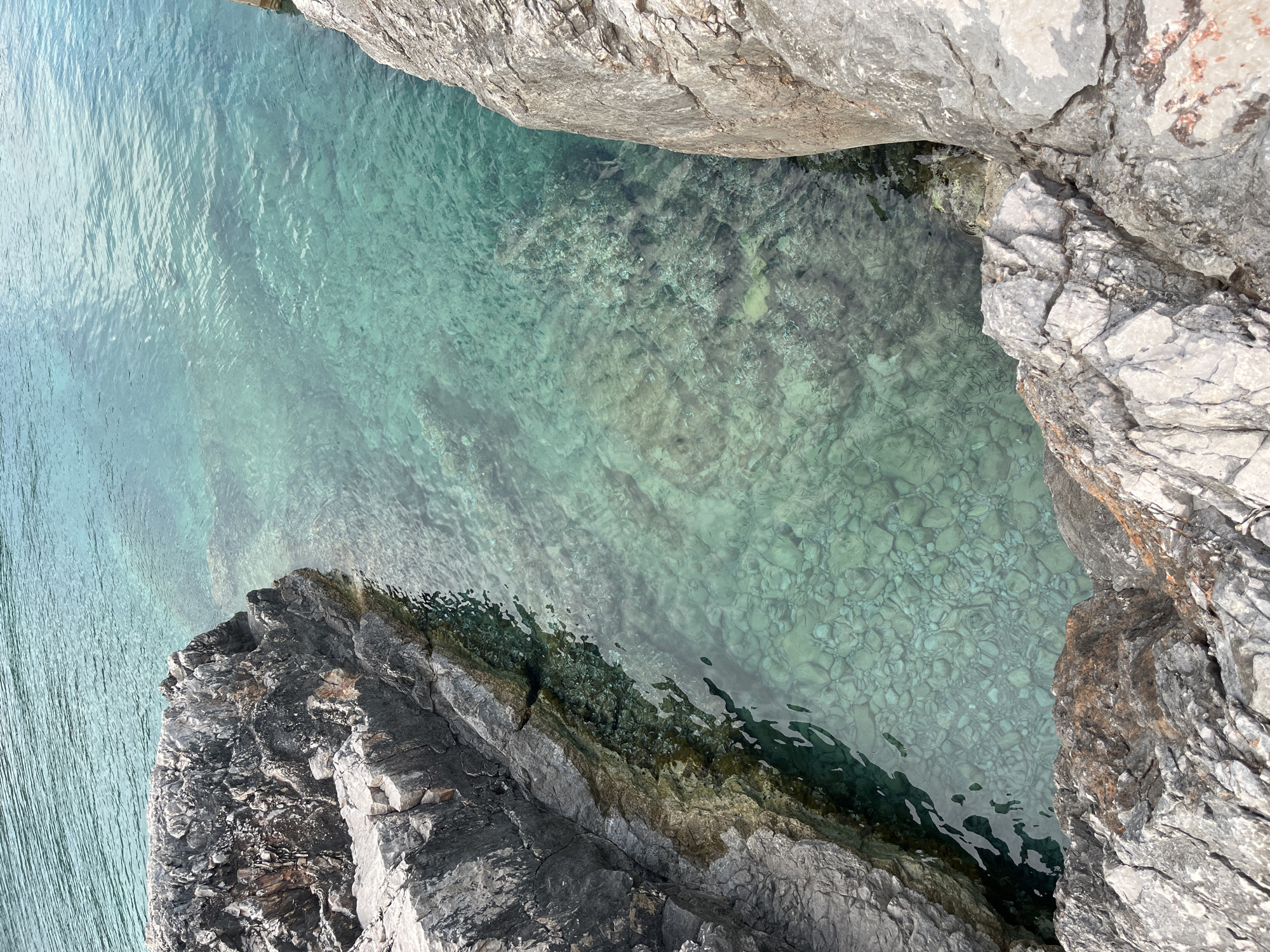 This media file shows a high-angle view of a rocky coastline and clear turquoise water. The water is shallow enough to see the rocky seabed beneath the surface, with pebbles and larger rocks visible. The water has gentle ripples, suggesting a calm day.
The foreground is dominated by dark, jagged rocks that form the edge of the coastline. These rocks are rough and textured, with some areas appearing wet. Further out, larger, lighter-colored rocks frame the scene, one of which is seen from above, its surface rough and weathered.
The overall scene is one of natural beauty and tranquility. The bright sunlight, indicated by the clear water and the bright illumination of the rocks, suggests it is likely daytime, possibly midday or afternoon. The location appears to be a secluded coastal area, possibly a cove or a bay, with no signs of human activity or structures visible. The environment evokes a sense of peacefulness and the raw beauty of nature. There is no visible text in the image.
