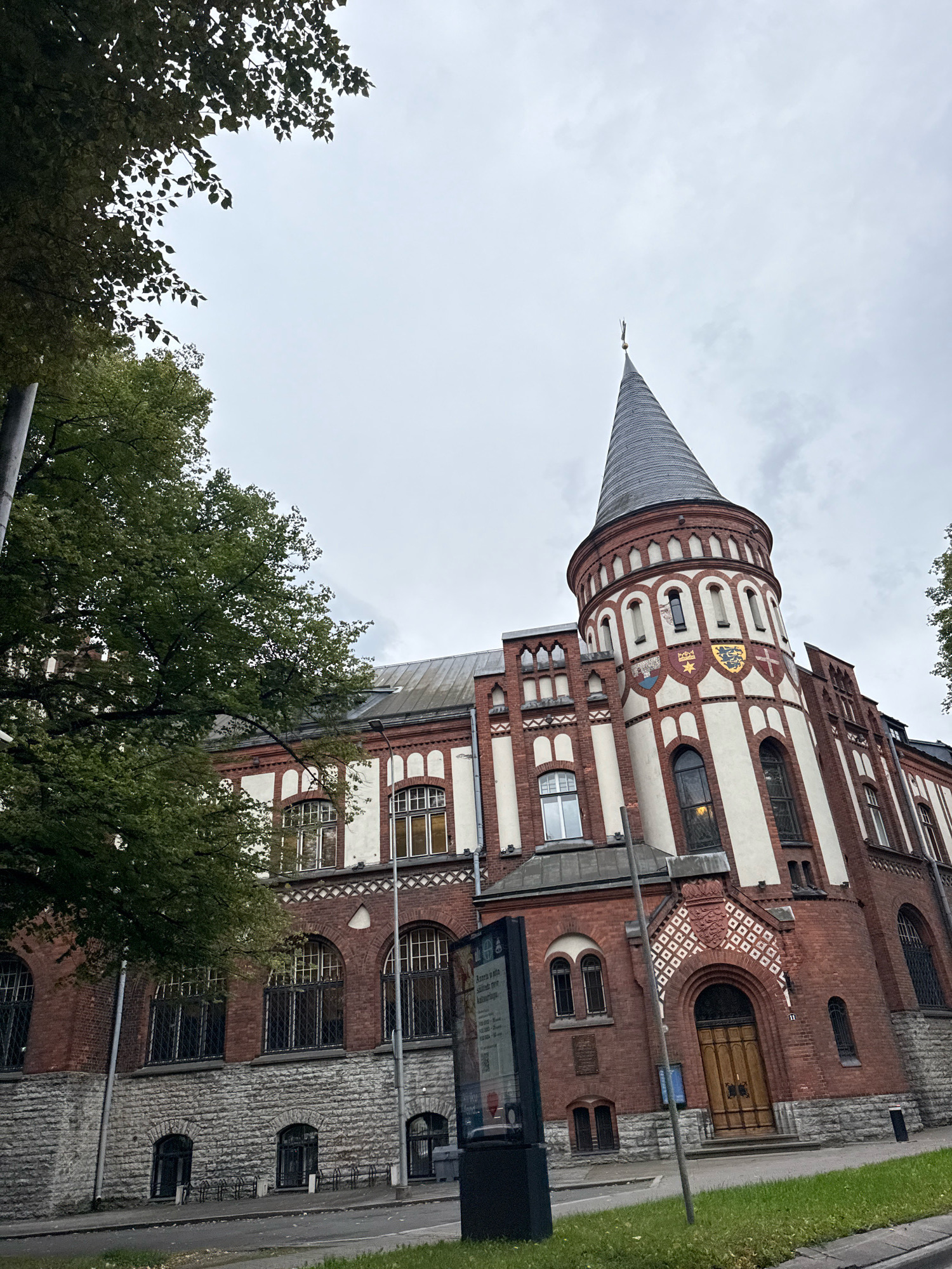 This is a low-angle, medium shot of the exterior of the Estonian Maritime Museum (Eesti Meremuuseum) located in Tallinn, Estonia. The building, constructed primarily of red brick with white accents, features a prominent conical tower with a gray roof and a golden finial. The tower's facade is decorated with arched windows and coats of arms.
To the left of the building, a large tree with green foliage partially obscures the view of the facade. The sky is overcast and gray. In the foreground, a street runs alongside the building, with a sidewalk and some grass visible. A modern information display stands on the sidewalk to the left of the entrance. The entrance itself is an arched wooden door set into the brick facade. Bicycle racks are visible in front of some of the ground-floor windows. The overall scene suggests an urban environment on a cloudy day.