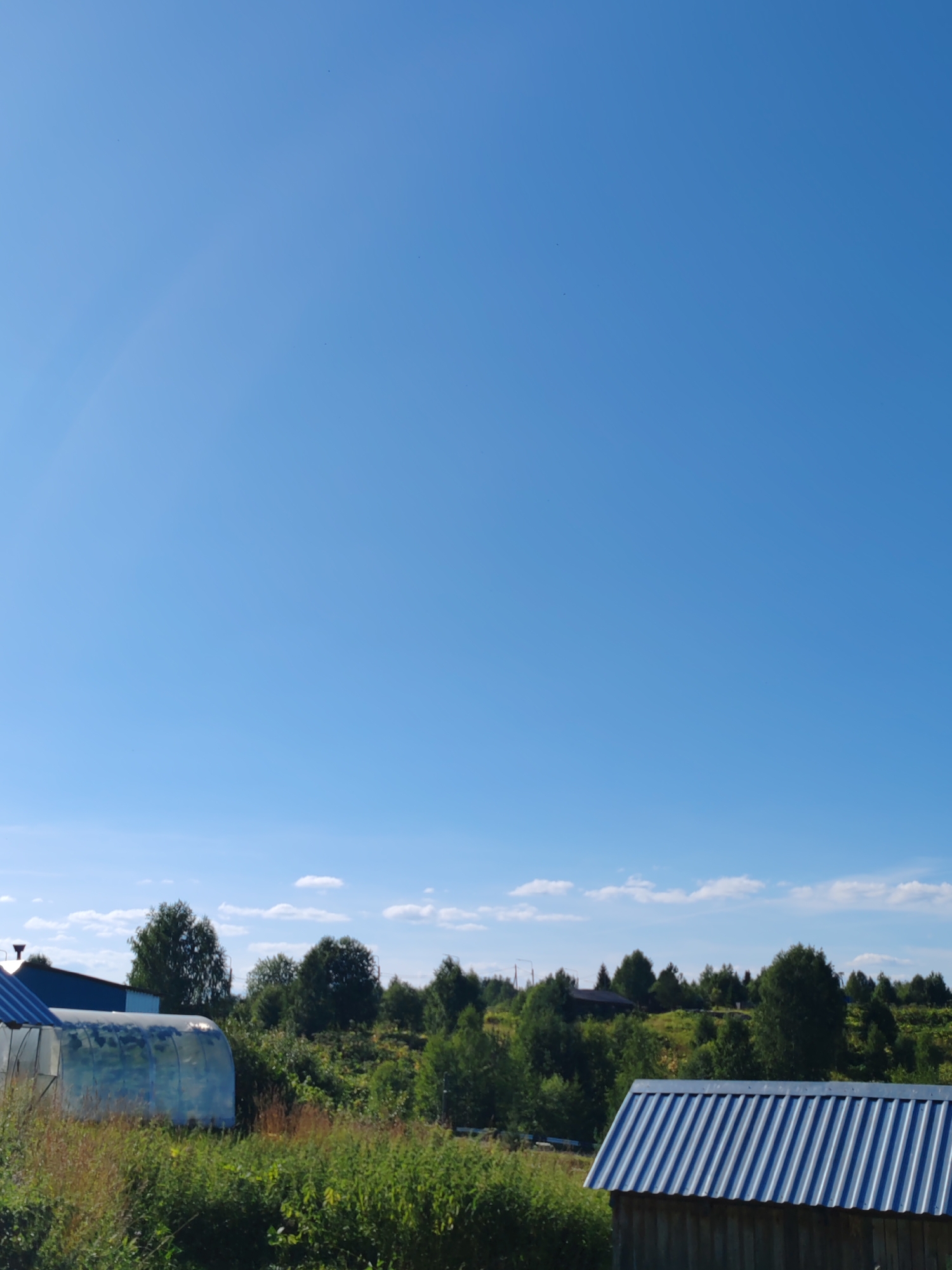 The image depicts a rural landscape under a clear blue sky, with scattered clouds on the horizon. In the foreground, to the left, a greenhouse with a transparent cover is visible, partially obscured by lush green vegetation. To the right, the corrugated metal roof of a wooden structure is prominent. The middle ground is characterized by a gentle slope covered in dense green trees and shrubs. Further in the distance, several houses with dark roofs and trees are scattered across the landscape. The overall scene suggests a serene summer day in a village or countryside setting, likely in the city of Chukhlem, Russia, given the provided context, characterized by agricultural structures and abundant greenery. There are no people or animals visible in the image.