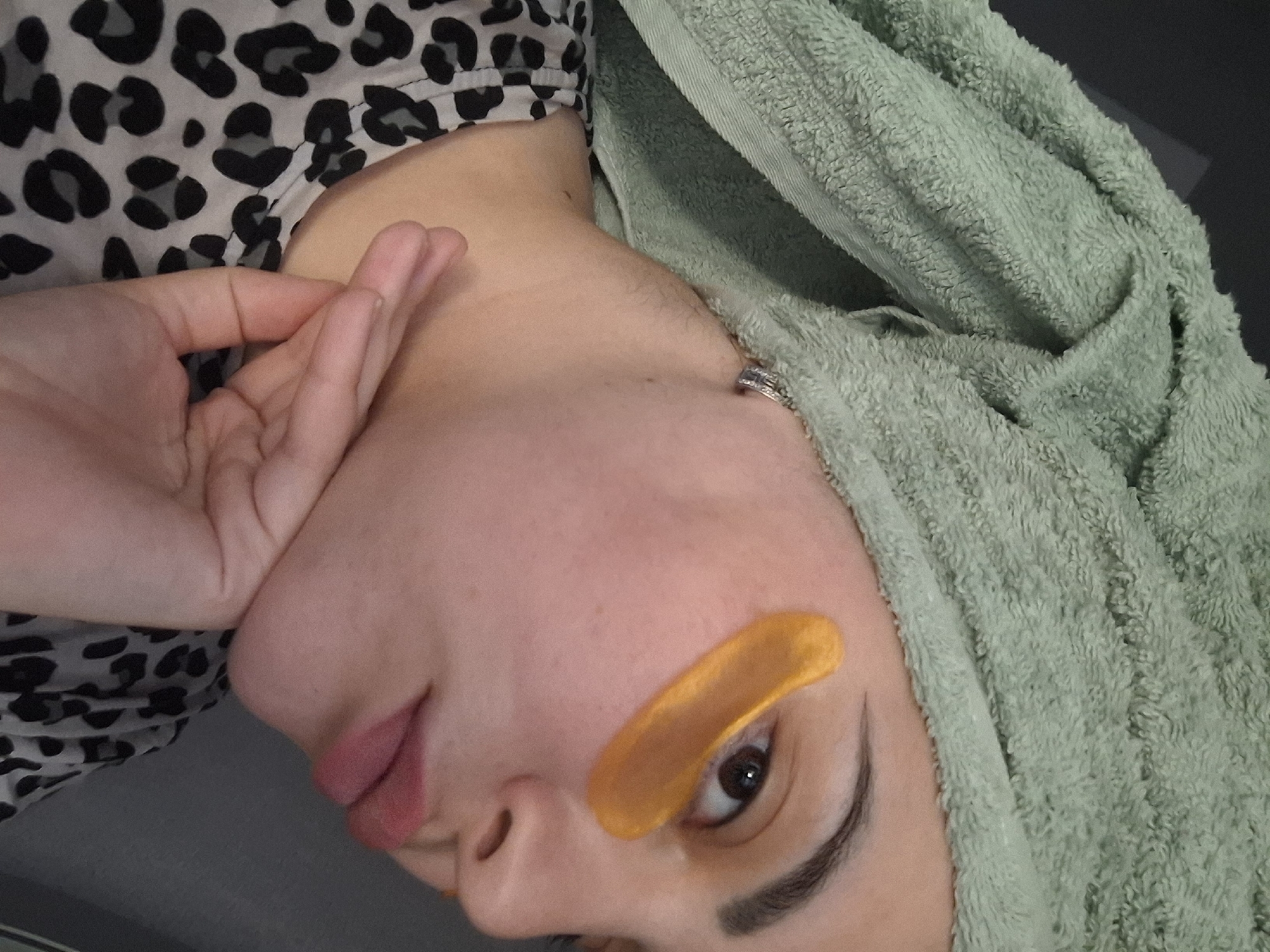 A young woman's face is visible, turned sideways. She is wearing an animal print top and has a light green towel wrapped around her head. An orange under-eye mask is present below her left eye. Her right hand is near her face, and she is wearing a ring on her finger. The location in the city of Амзя, Russia is not verifiable from the image.